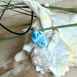 Larimar Freeform-Anhänger auf schwarzer Lederkette. Beidseitig tragbarer Trommelstein, ca. 2 x 1,8 cm groß, mit Silberöse. Der seltene, kostbare Larimar zeigt lebendige Blau- und Weißnuancen wie karibisches Lagunenwasser. Lederkette 45 cm mit Edelstahl-Karabiner und 5 cm Edelstahl-Verlängerung, fotografiert vor einer großen Muschel im maritimen Stil.