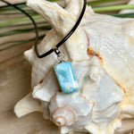 Larimar Freeform Anhänger an schwarzer Lederkette. Natürlicher, rechteckiger Larimar-Stein (ca. 1,5 × 1 cm) in Türkis-Blau mit weißen Strukturen, eingefasst in eine rhodinierte Silberöse. Beidseitig tragbar, an 45 cm Lederkette mit Edelstahl-Karabinerverschluss und 5 cm Verlängerung. Maritimes, zurückhaltend elegantes Schmuckstück mit der Optik karibischen Meerwassers.