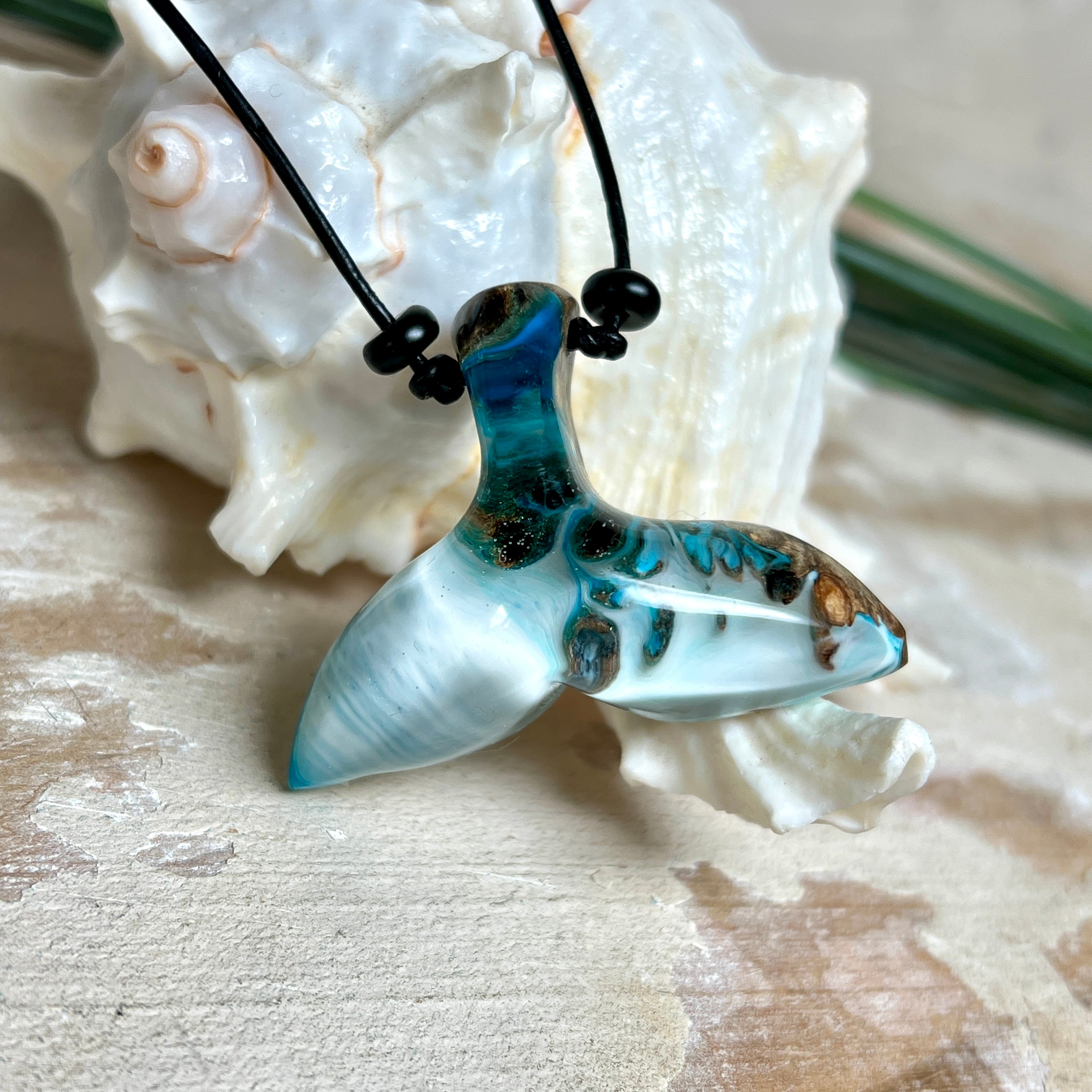 Handgefertigter Walflossen-Anhänger „Flossenklang“ aus Maserholz und türkis-blau-grau-weiß marmoriertem Epoxidharz an verstellbarem Lederband. Maritime Kette als Unikatschmuck für Meeresliebhaber und Wal-Fans, hochwertig poliert, präsentiert vor Muscheldeko und in Schmuckbox von ZeitlosSchmuckDesign.