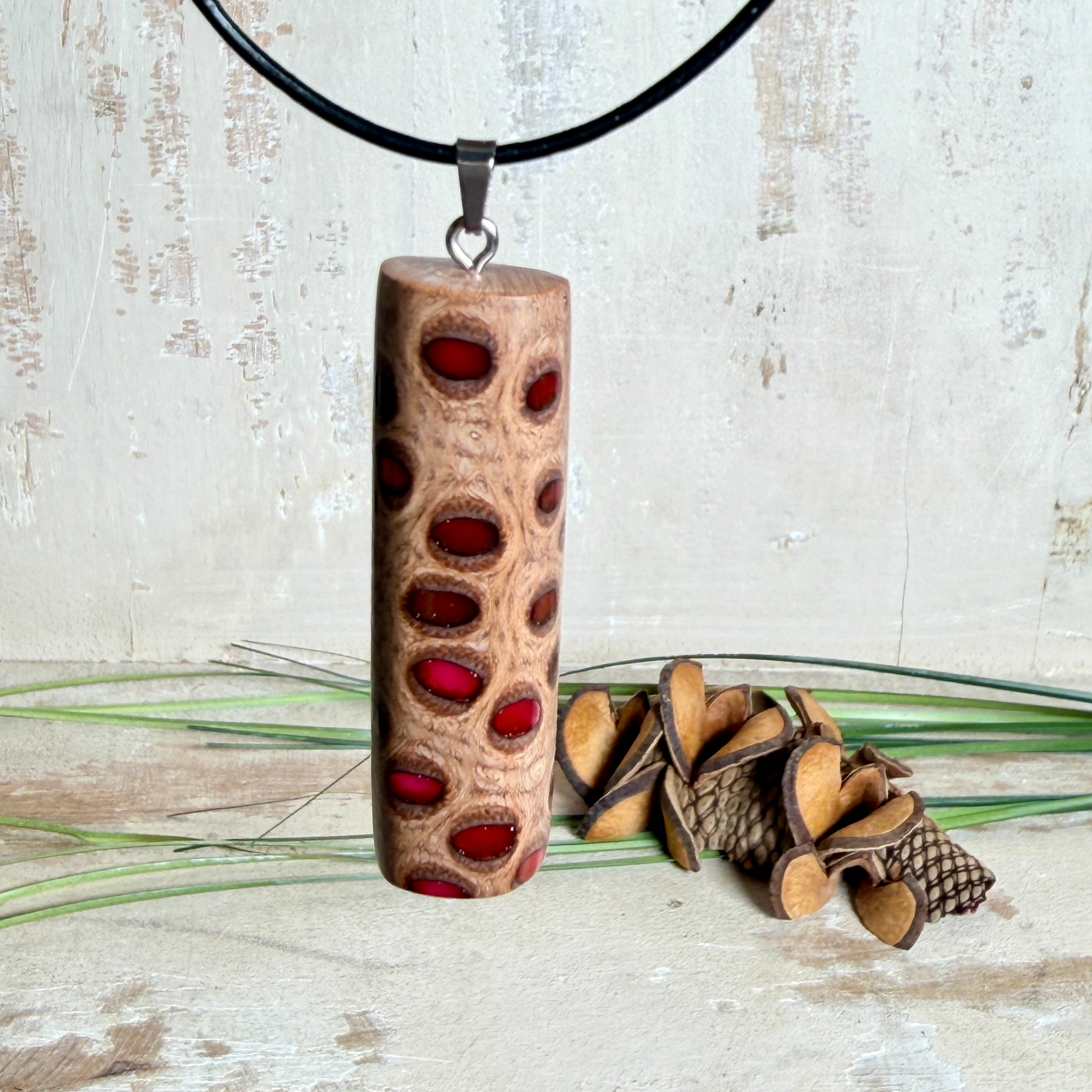 Handgefertigter Halsanhänger aus echtem Banksiazapfen, veredelt mit rotem Harz in den natürlichen Kammern; ca. 5,5 × 1,6 cm. Mit Anhängerschlaufe aus Edelstahl an schwarzer Lederkette (45 cm + 5 cm Verlängerung) mit Edelstahl-Karabiner. Detailaufnahme bzw. Tragebild vor neutralem, natürlichem Hintergrund.