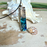 Rechteckiger Stabanhänger aus Maserholz und glasklarem Epoxidharz in tiefen Blau- und Türkistönen mit organischer Lagunen/Riff-Optik. Hochglanzpoliert, ca. 5,3 × 1,2 cm. Der Anhänger hängt an einer schwarzen Lederkette mit Edelstahl-Karabiner und 5-cm-Verlängerung (45 + 5 cm); die Anhängerschlaufe ist ebenfalls aus Edelstahl. Handgefertigtes Unikat, Lieferung in edler Schmuckbox – maritimer Look.