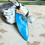 Handgefertigter Surfbrett-Anhänger aus warm gemasertem Olivenholz und Epoxidharz in Blau und Weiß, das an Meereswellen erinnert. Längliche, vorne polierte Form, ca. 6,2 × 2 cm, an verstellbarem schwarzem Lederband mit Knoten. Maritimer Unikatschmuck für Damen und Herren, dekorativ mit Stein und Muschel arrangiert.