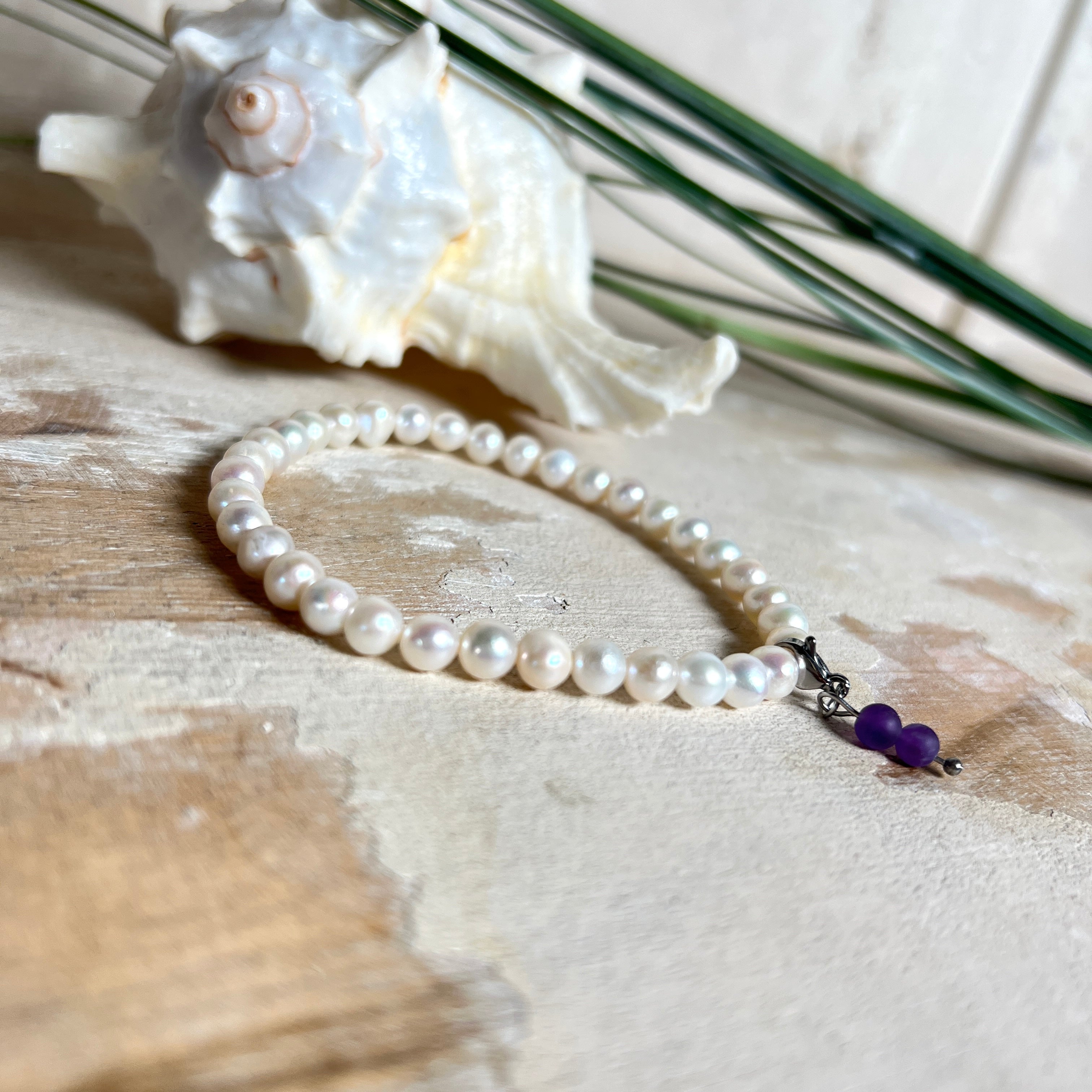 Handgefertigtes Armband „Sea Harmony“ mit schimmernden Süßwasserperlen (Ø ca. 6–7 mm), jede Perle ein Unikat. Auf elastischem Band gefädelt, mit einem filigranen Charmanhänger aus  Amethyst-Perlen, befestigt an einem robusten Edelstahl-Karabiner. Ein Schmuckstück voller Eleganz, maritimer Harmonie und symbolischer Tiefe – ideal als Geschenk oder als stilvolles Accessoire.