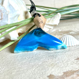 Handgefertigter Walflossen-Anhänger aus Olivenholz und glänzendem Harz in Blau-Grün-Weiß – maritimer Schmuck „Flossenklang“. Der Anhänger erinnert an die Form einer Walflosse und vereint die natürliche Maserung des Holzes mit dem schimmernden Farbspiel des Harzes. Mit verstellbarem schwarzen Lederband und Spacerperle. Ideal für Meeresliebhaber und Naturliebhaber – jedes Stück ein Unikat.