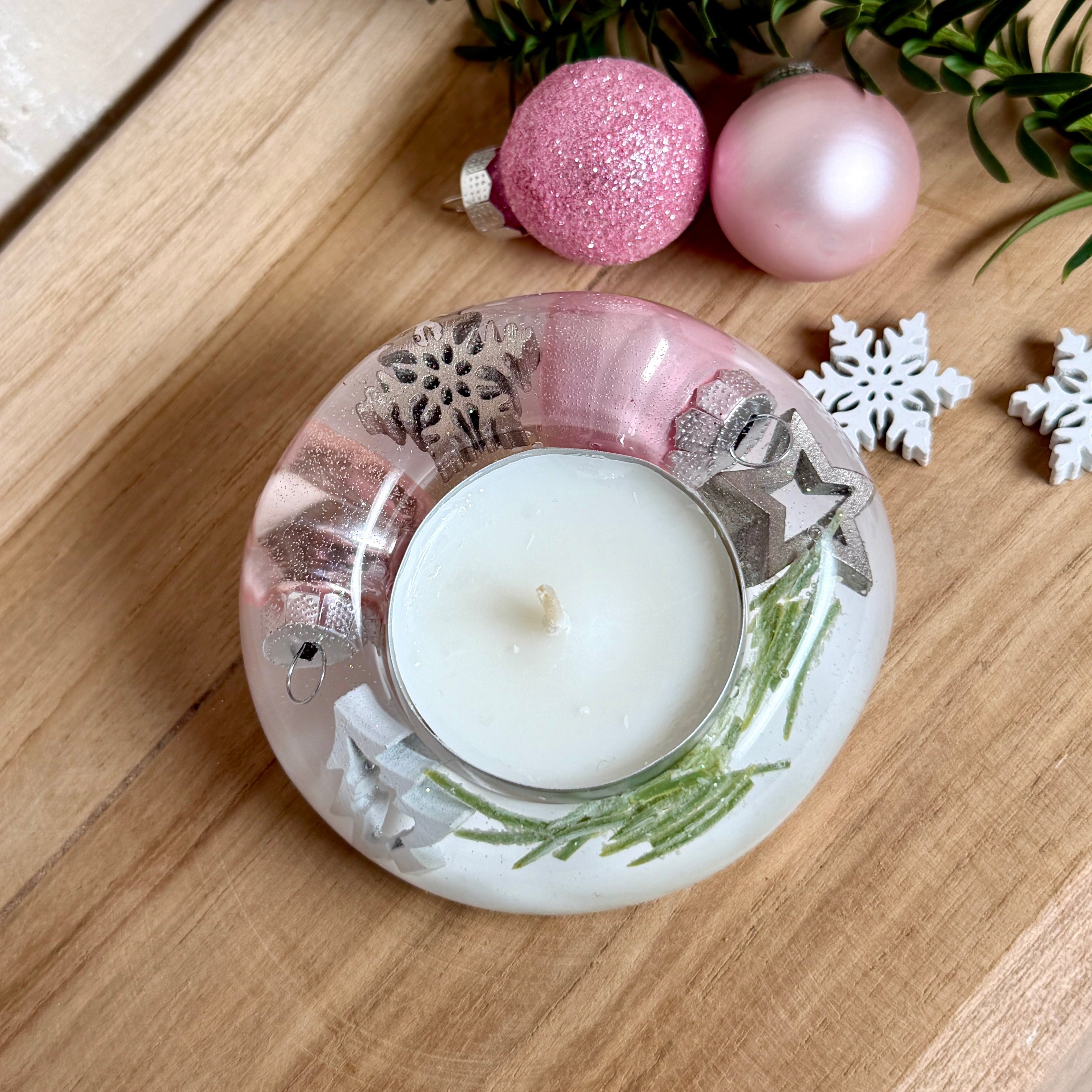 Runder Teelichthalter aus klarem Epoxidharz, Durchmesser ca. 7 cm, Höhe ca. 4 cm. Im Harz sind rosa Weihnachtskugeln, ein Stern, eine Schneeflocke, ein Tannenbaum, künstliche Tannennadeln und feiner Glitzer eingegossen. Vertiefung für ein Teelicht in der Mitte, Unterseite mit vier Korkpads als Möbelschoner. Fotografiert auf Holz mit Tannenzweig und rosa Kugeln als weihnachtliche Dekoration