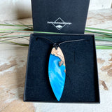 Handgefertigter Surfbrett-Anhänger aus warm gemasertem Olivenholz und Epoxidharz in Blau und Weiß, das an Meereswellen erinnert. Längliche, vorne polierte Form, ca. 6,2 × 2 cm, an verstellbarem schwarzem Lederband mit Knoten. Maritimer Unikatschmuck für Damen und Herren, dekorativ mit Stein und Muschel arrangiert.