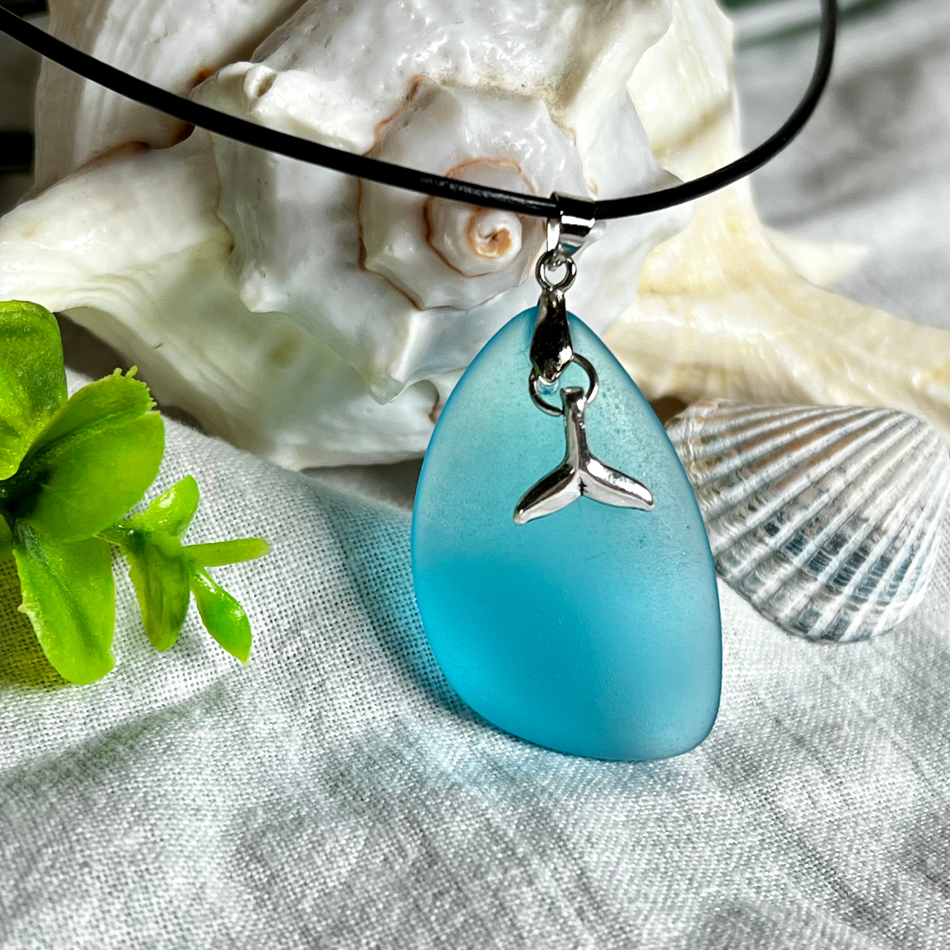 Edle Halskette „Blue Essence“ mit handgefertigtem türkisfarbenem Seeglas-Anhänger und Edelstahl-Flosse. Die Kette besteht aus schwarzer Lederkordel mit Karabinerverschluss und Verlängerung aus Edelstahl. Das matte Seeglas reflektiert Licht wie Meereswasser – ideal für Strandliebhaber und Naturliebhaber.