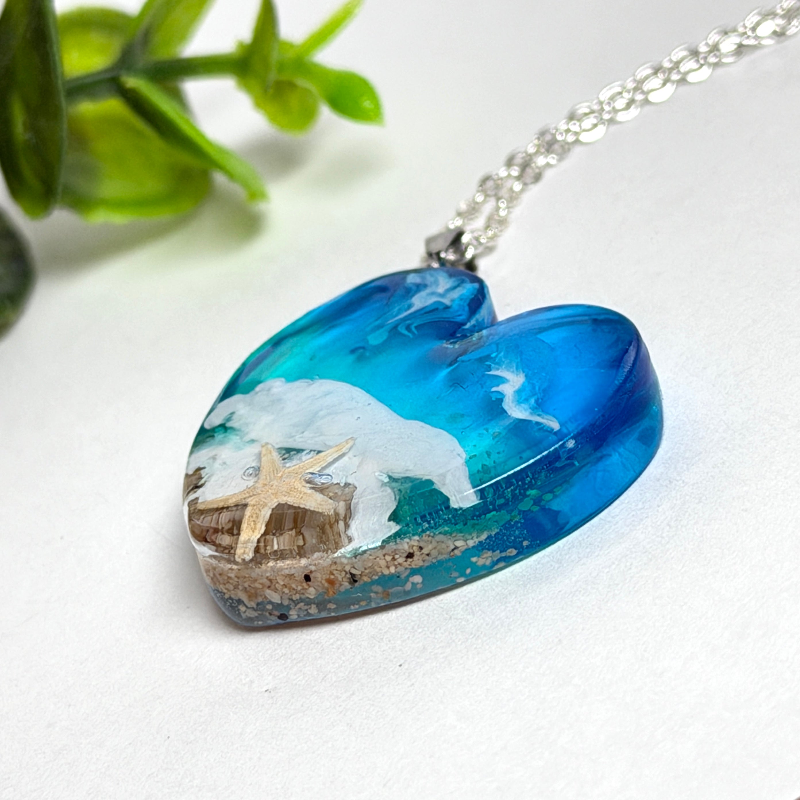 Ein herzförmiger Anhänger mit einer ganzen Ozeanwelt darin: Welle, Sand und Seestern in leuchtendem Blau und Türkis – handgegossen, einzigartig, unvergesslich. Mit versilberter Kette (46 cm) und Edelstahl-Karabiner.