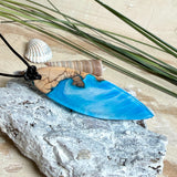 Handgefertigter Surfbrett-Anhänger aus warm gemasertem Olivenholz und Epoxidharz in Blau und Weiß, das an Meereswellen erinnert. Längliche, vorne polierte Form, ca. 6,2 × 2 cm, an verstellbarem schwarzem Lederband mit Knoten. Maritimer Unikatschmuck für Damen und Herren, dekorativ mit Stein und Muschel arrangiert.