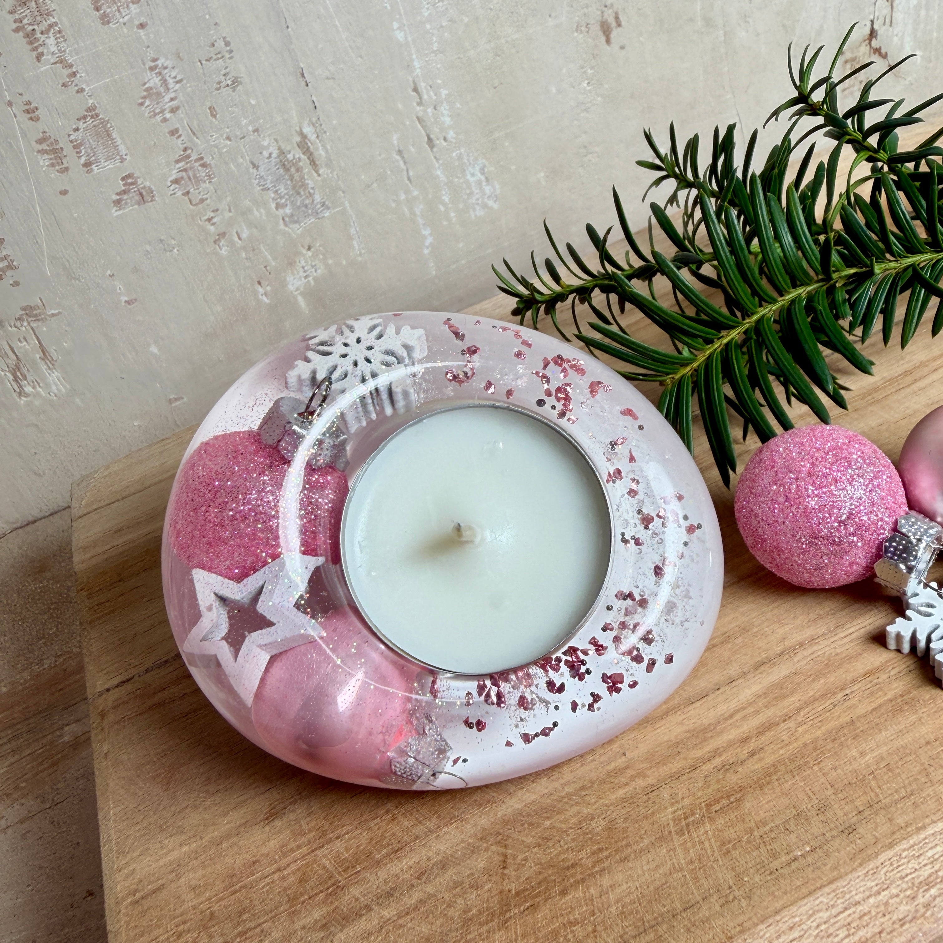 Rosa Teelichthalter aus klarem Epoxidharz, ca. 8 × 6,5 cm, Höhe ca. 4 cm. Im Harz sind kleine rosa Weihnachtskugeln, ein Stern, eine Schneeflocke und rosa Granulat eingegossen. Ovaler Halter für ein Teelicht, Unterseite mit vier runden Korkpads als Möbelschoner. Fotografiert auf Holz mit Tannenzweig und rosa Kugeln als weihnachtlicher Dekohintergrund.