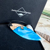 Handgefertigter Surfbrett-Anhänger aus warm gemasertem Olivenholz und Epoxidharz in Blau und Weiß, das an Meereswellen erinnert. Längliche, vorne polierte Form, ca. 6,2 × 2 cm, an verstellbarem schwarzem Lederband mit Knoten. Maritimer Unikatschmuck für Damen und Herren, dekorativ mit Stein und Muschel arrangiert.