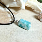 Larimar Freeform Anhänger an schwarzer Lederkette. Natürlicher, rechteckiger Larimar-Stein (ca. 1,5 × 1 cm) in Türkis-Blau mit weißen Strukturen, eingefasst in eine rhodinierte Silberöse. Beidseitig tragbar, an 45 cm Lederkette mit Edelstahl-Karabinerverschluss und 5 cm Verlängerung. Maritimes, zurückhaltend elegantes Schmuckstück mit der Optik karibischen Meerwassers.