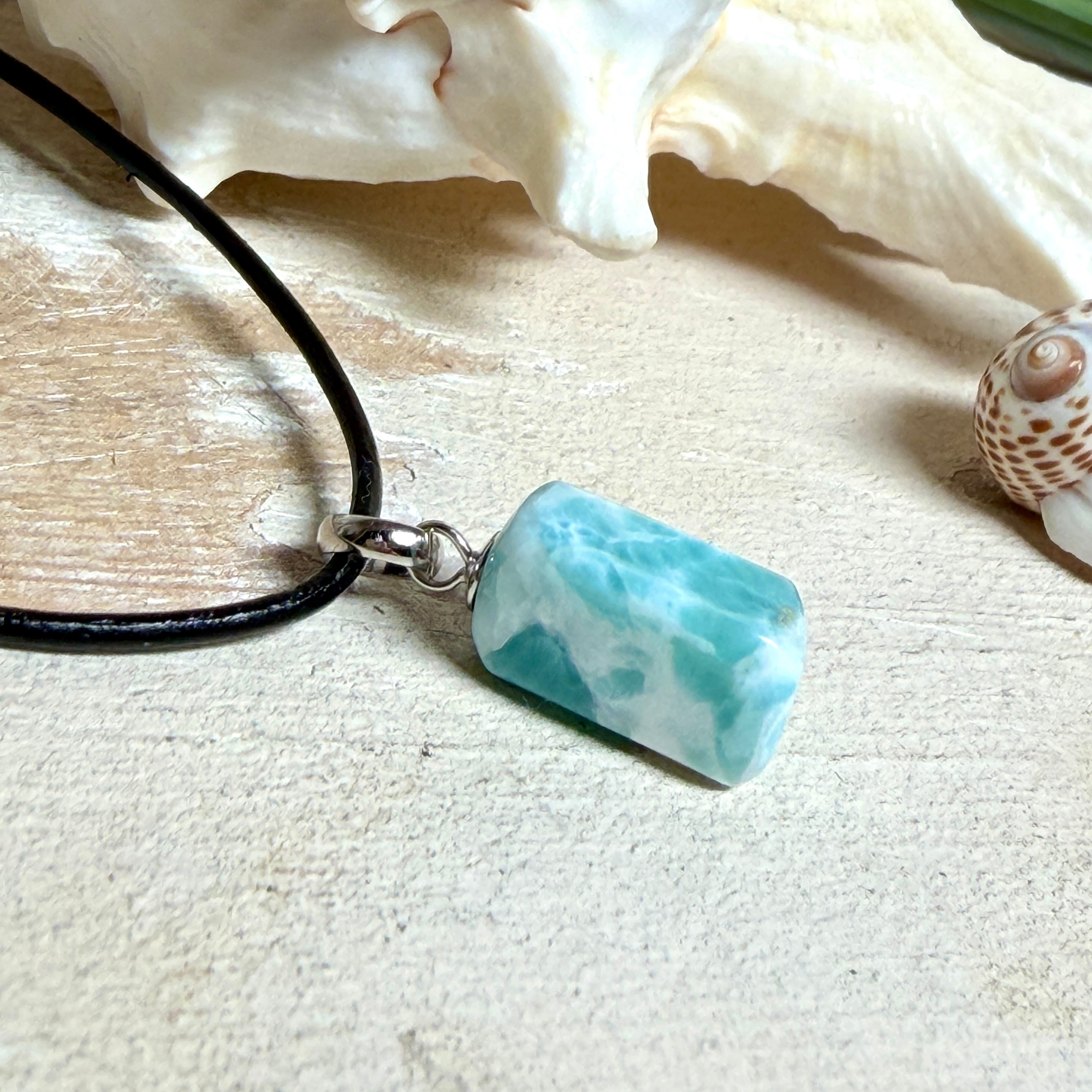 Larimar Freeform Anhänger an schwarzer Lederkette. Natürlicher, rechteckiger Larimar-Stein (ca. 1,5 × 1 cm) in Türkis-Blau mit weißen Strukturen, eingefasst in eine rhodinierte Silberöse. Beidseitig tragbar, an 45 cm Lederkette mit Edelstahl-Karabinerverschluss und 5 cm Verlängerung. Maritimes, zurückhaltend elegantes Schmuckstück mit der Optik karibischen Meerwassers.