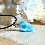 Larimar Freeform-Anhänger auf schwarzer Lederkette. Beidseitig tragbarer Trommelstein, ca. 2 x 1,8 cm groß, mit Silberöse. Der seltene, kostbare Larimar zeigt lebendige Blau- und Weißnuancen wie karibisches Lagunenwasser. Lederkette 45 cm mit Edelstahl-Karabiner und 5 cm Edelstahl-Verlängerung, fotografiert vor einer großen Muschel im maritimen Stil.