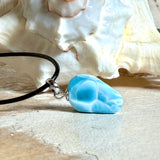 Larimar Freeform-Anhänger auf schwarzer Lederkette. Beidseitig tragbarer Trommelstein, ca. 2 x 1,8 cm groß, mit Silberöse. Der seltene, kostbare Larimar zeigt lebendige Blau- und Weißnuancen wie karibisches Lagunenwasser. Lederkette 45 cm mit Edelstahl-Karabiner und 5 cm Edelstahl-Verlängerung, fotografiert vor einer großen Muschel im maritimen Stil.