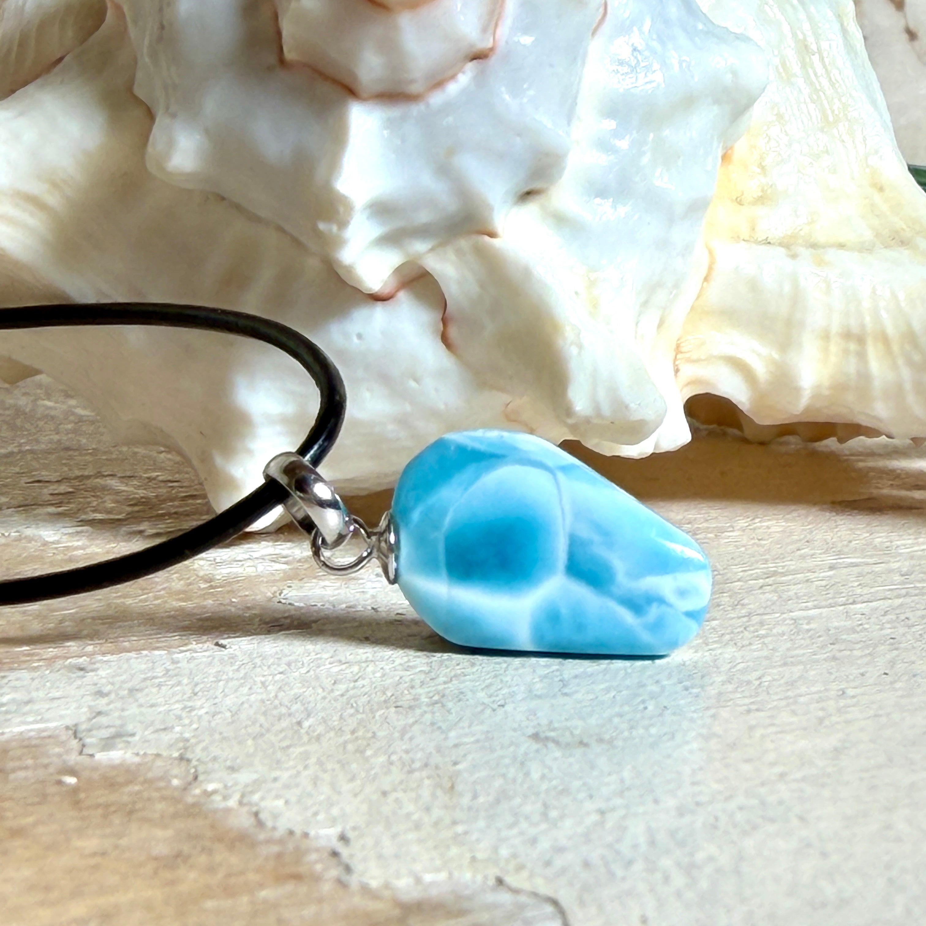 Larimar Freeform-Anhänger auf schwarzer Lederkette. Beidseitig tragbarer Trommelstein, ca. 2 x 1,8 cm groß, mit Silberöse. Der seltene, kostbare Larimar zeigt lebendige Blau- und Weißnuancen wie karibisches Lagunenwasser. Lederkette 45 cm mit Edelstahl-Karabiner und 5 cm Edelstahl-Verlängerung, fotografiert vor einer großen Muschel im maritimen Stil.