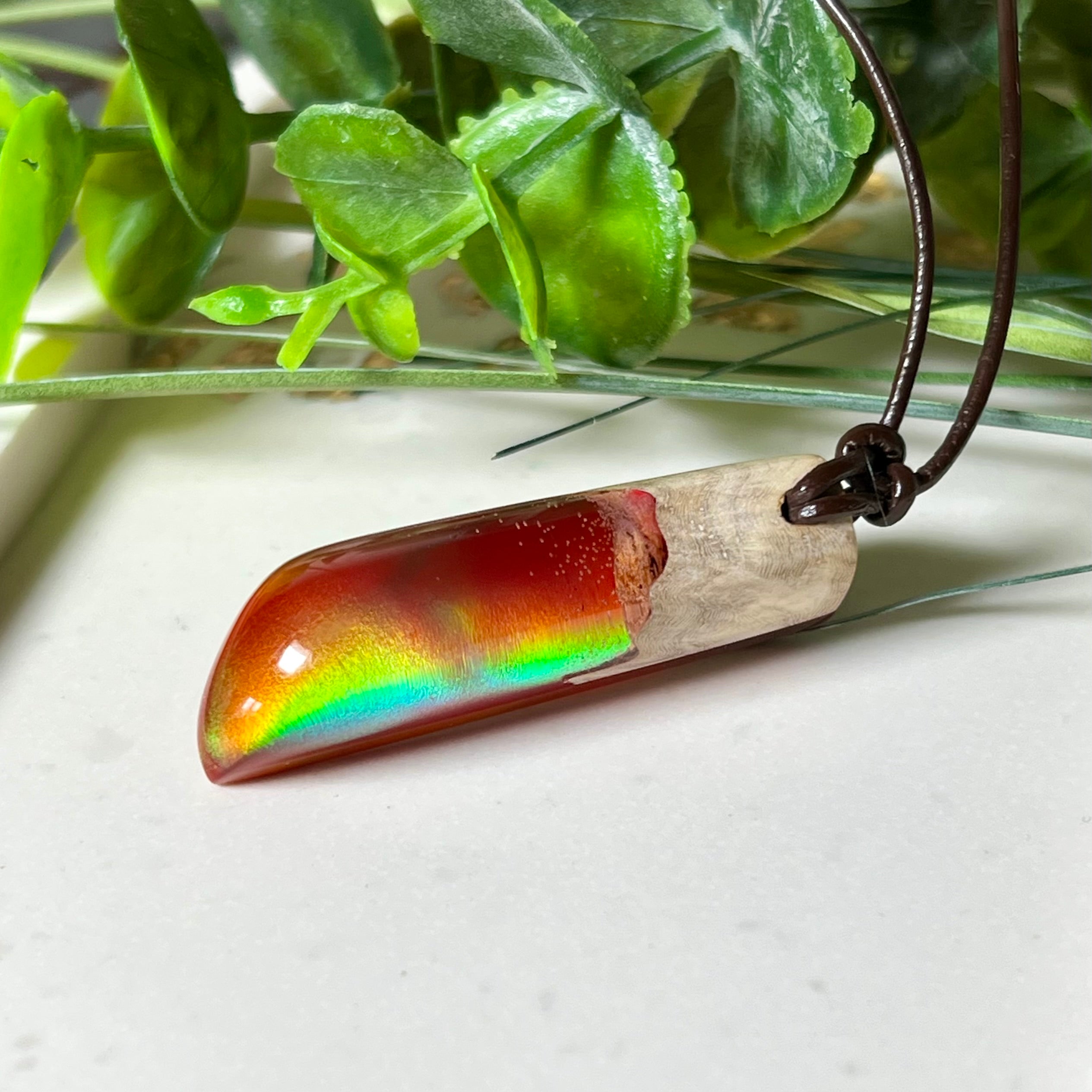 Handgefertigter Hei Toki Anhänger aus Goldfield-Maserholz und rubinrotem Labor-Opal, eingebettet in glänzendes Epoxidharz. Je nach Lichteinfall schimmert das Harz in warmen Farben mit Regenbogeneffekt. Der Anhänger ist ca. 4,3 × 1,5 cm groß und wird an einem verstellbaren Lederband mit Holzperlen getragen. Symbolischer Unikatschmuck – ideal als Geschenk für besondere Menschen.