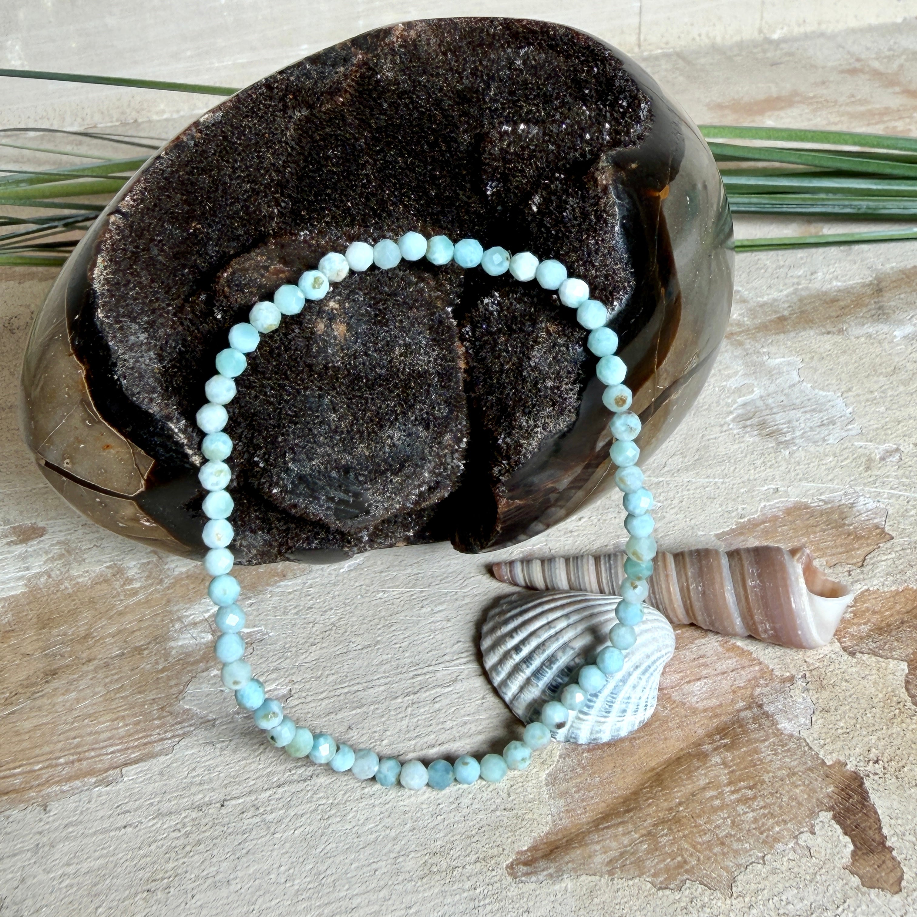 Zartes Larimar-Armband „Lagunenlicht“ mit facettierten 3 mm Perlen aus seltenem Atlantisstein auf Elastikfaden. Die kleinen Larimarsteine zeigen sanfte Blau- und Weißtöne wie Lagunenwasser. Innenumfang ca. 19 cm. Fotografiert auf Muscheln, einem dekorativen Stein und in einer schwarzen Schmuckbox von ZeitlosSchmuckDesign im maritimen Stil.
