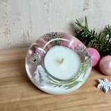 Runder Teelichthalter aus klarem Epoxidharz, Durchmesser ca. 7 cm, Höhe ca. 4 cm. Im Harz sind rosa Weihnachtskugeln, ein Stern, eine Schneeflocke, ein Tannenbaum, künstliche Tannennadeln und feiner Glitzer eingegossen. Vertiefung für ein Teelicht in der Mitte, Unterseite mit vier Korkpads als Möbelschoner. Fotografiert auf Holz mit Tannenzweig und rosa Kugeln als weihnachtliche Dekoration