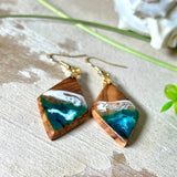 Handgefertigte Ohrringe „Ocean Diamond“ aus Olivenholz und Epoxidharz in Diamantform. Türkis-, Blau- und Weißtöne bilden ein wellenartiges Design, inspiriert vom Meer. Jeder Ohrring ist ein Unikat mit individueller Maserung. Vergoldete, nickelfreie und bleifreie Anhängerschlaufe, 925 Sterling Silber Ohrhaken. Maritimer Schmuck für Meerliebhaber und besondere Anlässe.