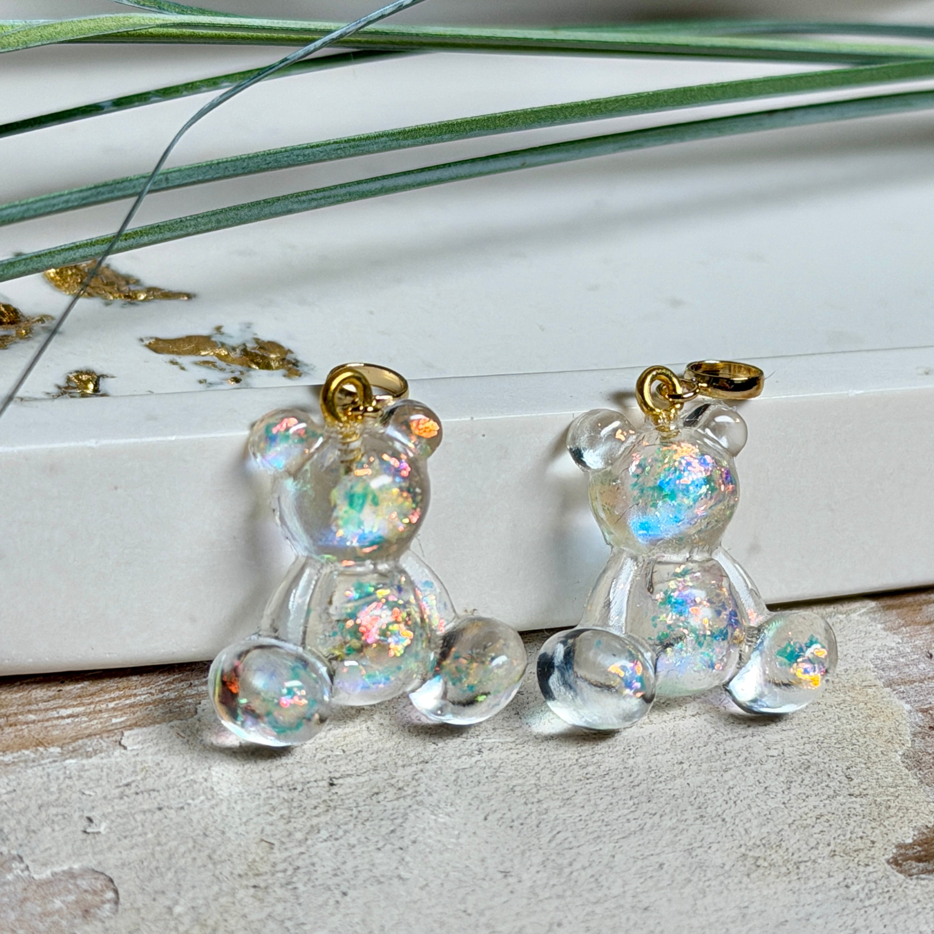 Kleiner Bärchen-Anhänger aus transparentem Harz mit irisierenden Flakes in Regenbogen-Schimmer, goldfarbene Anhängerschlaufe aus Edelstahl. Größe ca. 2,2 × 2,1 cm. Handgefertigtes Unikat; Flakes-Anordnung variiert, vereinzelt kleine Luftbläschen möglich.