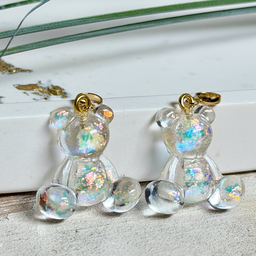 Kleiner Bärchen-Anhänger aus transparentem Harz mit irisierenden Flakes in Regenbogen-Schimmer, goldfarbene Anhängerschlaufe aus Edelstahl. Größe ca. 2,2 × 2,1 cm. Handgefertigtes Unikat; Flakes-Anordnung variiert, vereinzelt kleine Luftbläschen möglich.