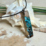 Rechteckiger Stabanhänger aus Maserholz und glasklarem Epoxidharz in tiefen Blau- und Türkistönen mit organischer Lagunen/Riff-Optik. Hochglanzpoliert, ca. 5,3 × 1,2 cm. Der Anhänger hängt an einer schwarzen Lederkette mit Edelstahl-Karabiner und 5-cm-Verlängerung (45 + 5 cm); die Anhängerschlaufe ist ebenfalls aus Edelstahl. Handgefertigtes Unikat, Lieferung in edler Schmuckbox – maritimer Look.