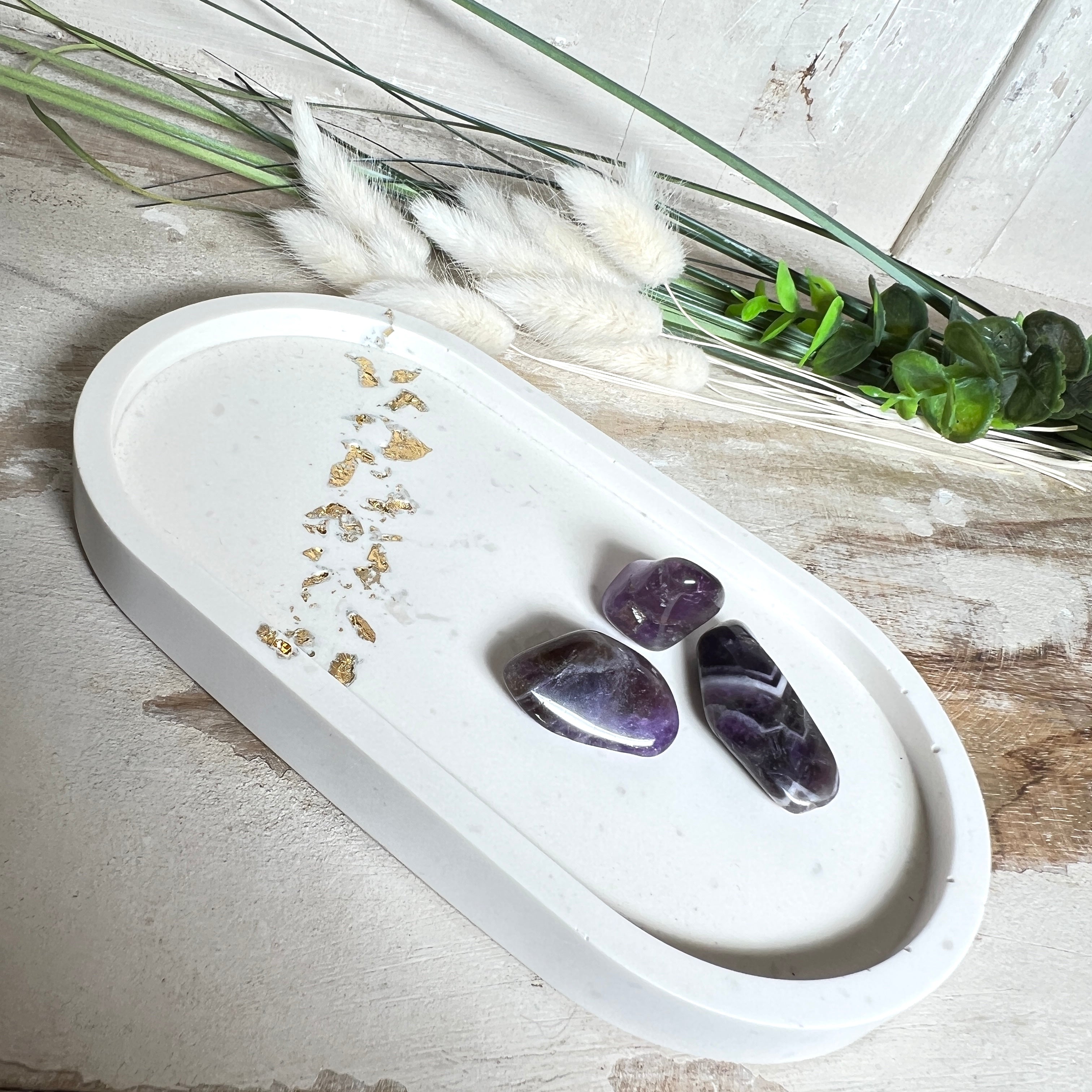 Handgefertigtes ovales Schmucktablett aus Jesmonite mit schimmernden Blattgold-Akzenten. Die Rückseite ist mit rutschfesten Korkpunkten ausgestattet. Ideal zur stilvollen Aufbewahrung von Schmuck, Schlüsseln oder kleinen Accessoires auf Kommoden und Nachttischen. Helles Design mit natürlicher Oberfläche und modernem Look. Unikat in feiner Handarbeit gefertigt – ohne Deko geliefert.