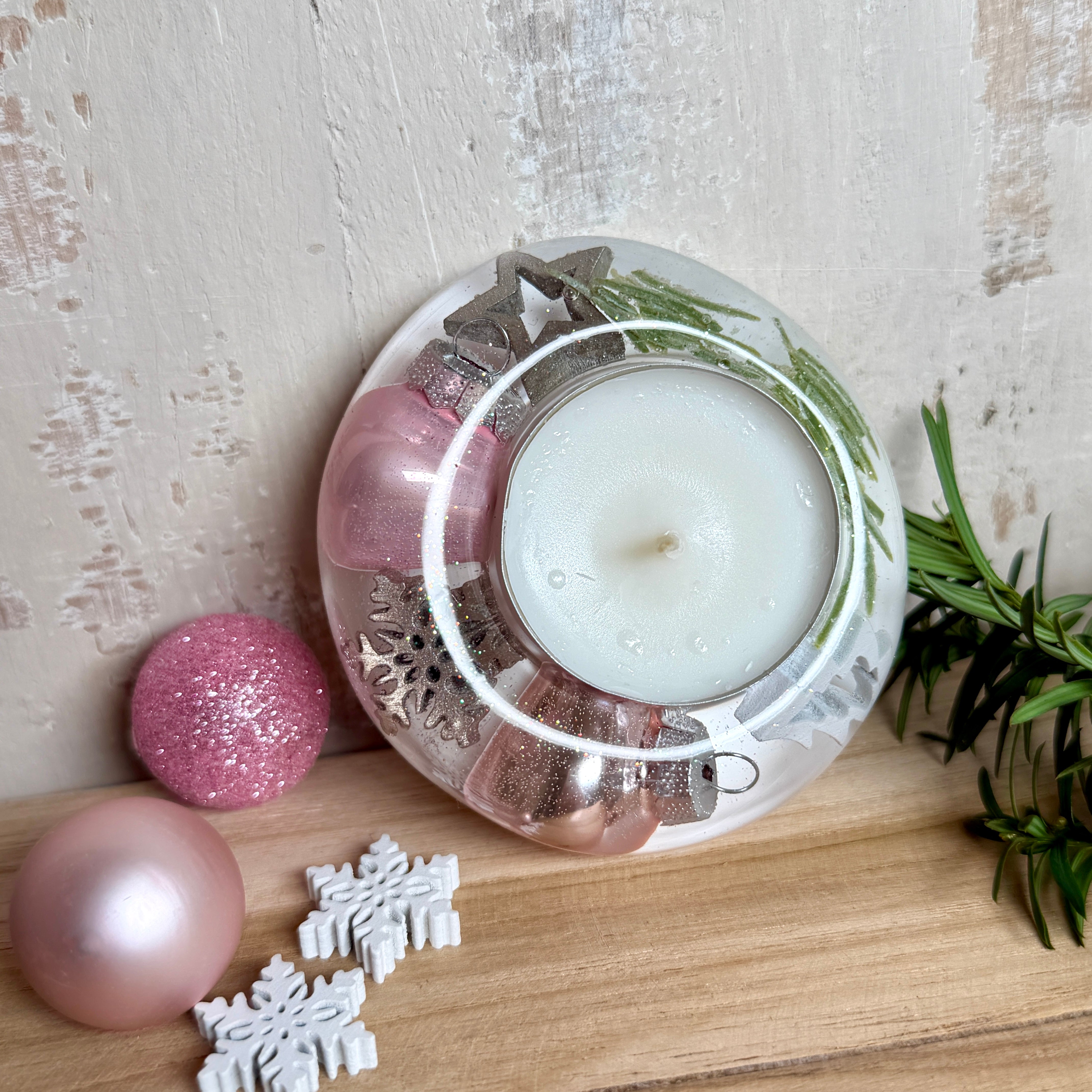 Runder Teelichthalter aus klarem Epoxidharz, Durchmesser ca. 7 cm, Höhe ca. 4 cm. Im Harz sind rosa Weihnachtskugeln, ein Stern, eine Schneeflocke, ein Tannenbaum, künstliche Tannennadeln und feiner Glitzer eingegossen. Vertiefung für ein Teelicht in der Mitte, Unterseite mit vier Korkpads als Möbelschoner. Fotografiert auf Holz mit Tannenzweig und rosa Kugeln als weihnachtliche Dekoration