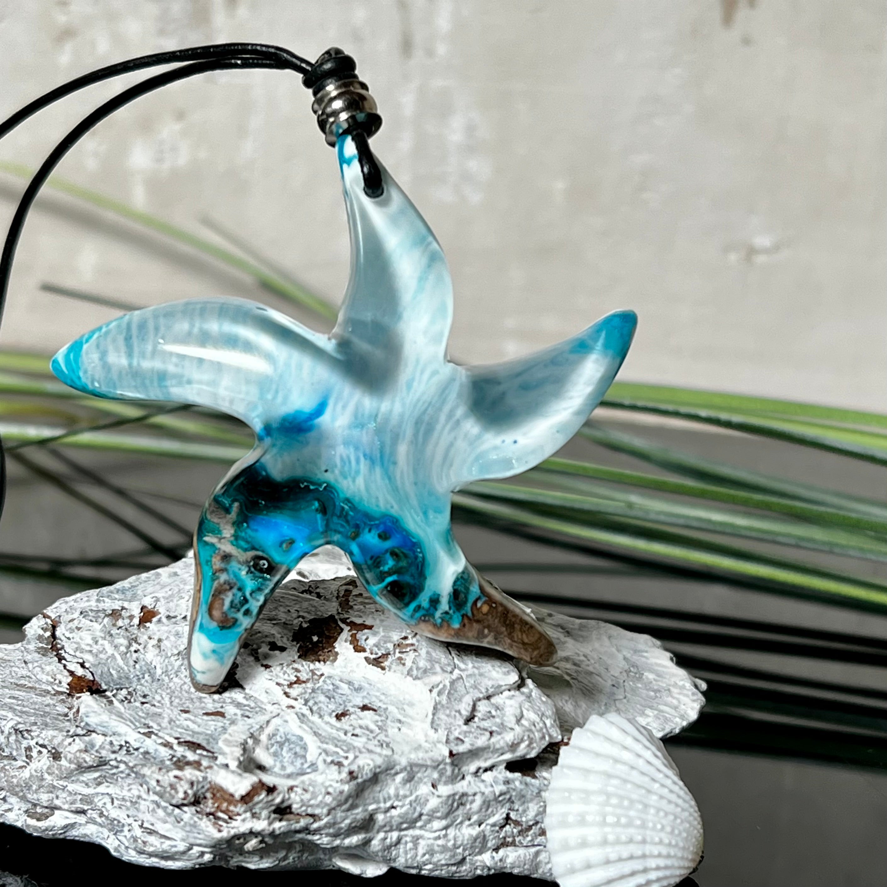 Handgefertigte Halskette in Seestern-Form mit Anhänger aus Naturholz und blau-weiß marmoriertem Epoxidharz. Das Schmuckstück ist einzigartig gestaltet und spiegelt maritimen Stil und Naturverbundenheit wider. Getragen an einem verstellbaren Lederband mit Holzperlen. Ideal für Meeresliebhaber und Naturfreunde.