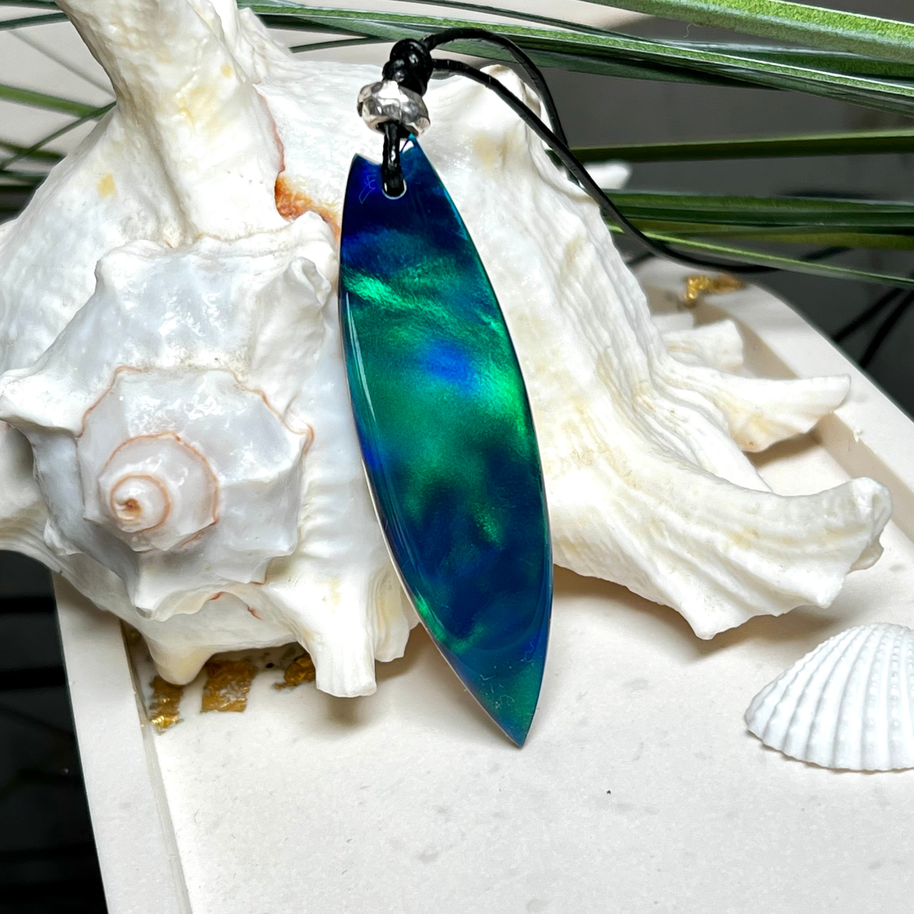 Beidseitig tragbarer Surfbrett-Anhänger aus Olivenholz, kombiniert mit klaren Epoxidharz in Blau-Grün. Eingearbeiteter synthetischer Opal erzeugt ein faszinierendes Farbspiel wie Nordlichter. Größe ca. 6 × 1,8 cm. Mit verstellbarem Lederband (26–45 cm), dekorativen Perlen & Schiebeknoten. Handgefertigt & einzigartig. Ein Schmuckstück mit Naturinspiration.
