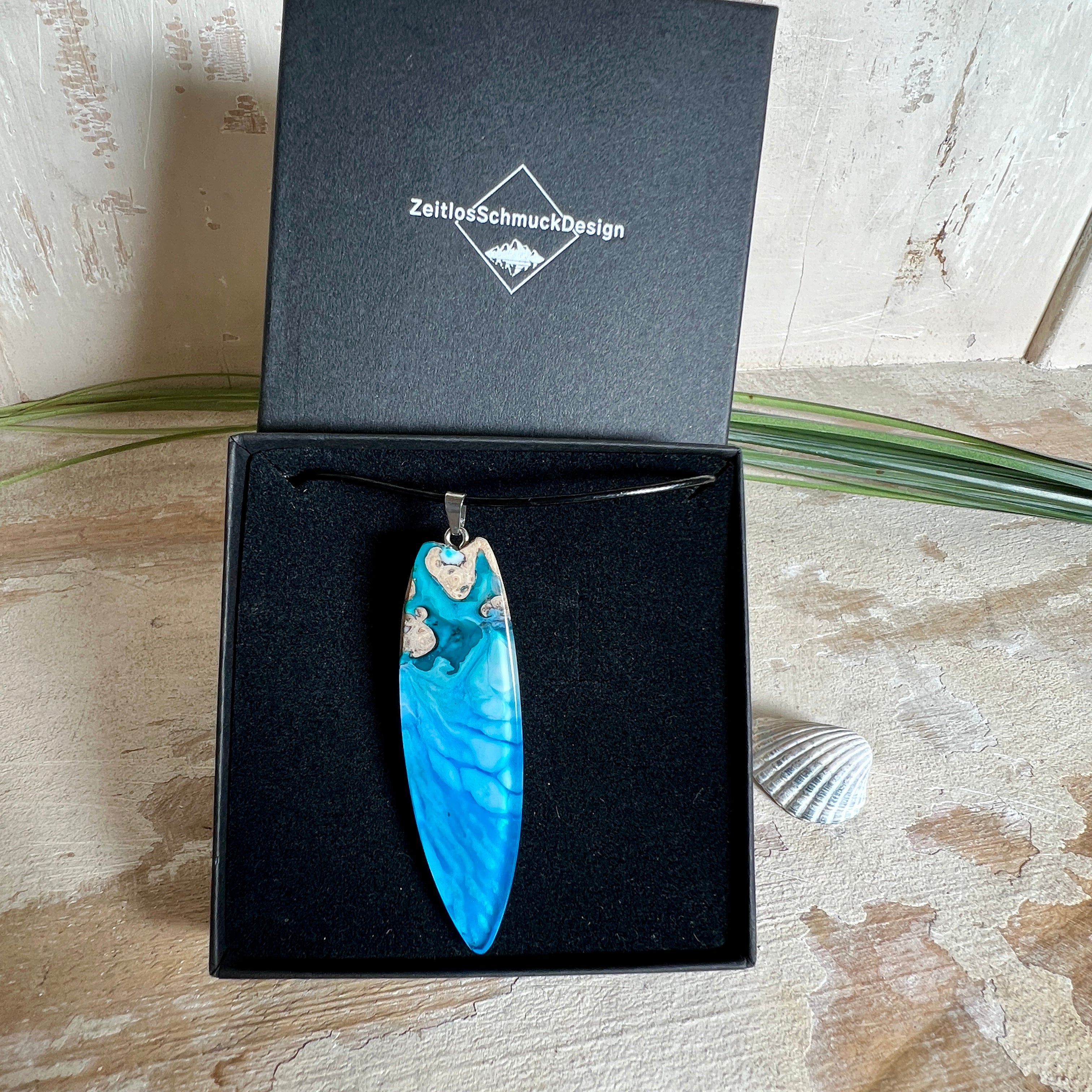Surfbrett-Anhänger aus Maserholz und blau-weißem Epoxidharz, hochglanzpoliert, ca. 6,5 × 2 cm. An schwarzer Lederkette mit Edelstahl-Karabiner und 5-cm-Verlängerung; Edelstahl-Öse. Im Tageslicht lagunenblau, unter Schwarzlicht fluoreszierend – ideal als Party-/Festival-Schmuck. Handgefertigtes Unikat, Lieferung in edler Schmuckbox.