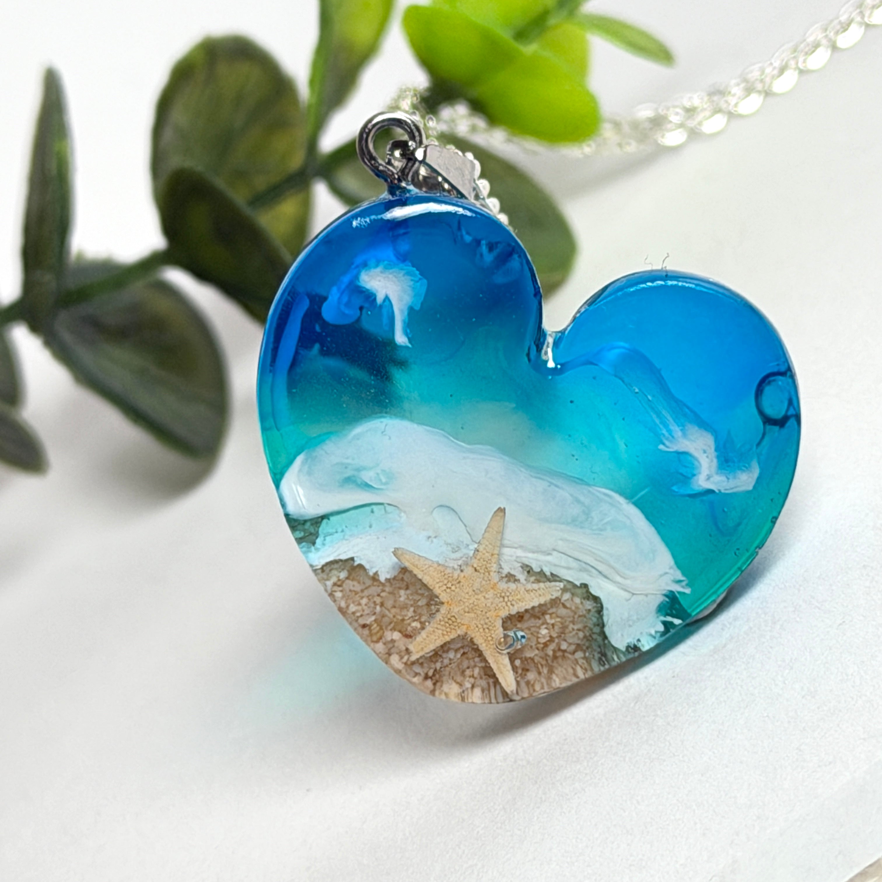 Ein herzförmiger Anhänger mit einer ganzen Ozeanwelt darin: Welle, Sand und Seestern in leuchtendem Blau und Türkis – handgegossen, einzigartig, unvergesslich. Mit versilberter Kette (46 cm) und Edelstahl-Karabiner.
