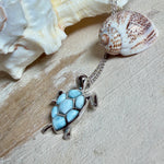 Feine Kette aus rhodiniertem 925 Silber mit Wasserschildkröten-Anhänger. Der ca. 25 x 18 mm große Anhänger zeigt eine Schildkröte, deren Panzer aus mehreren hellblauen Larimar-Inlays besteht. Seltener, kostbarer Larimar mit typischen wolkigen Strukturen, an einer ca. 45 cm langen, 1 mm starken, auf Platin rhodinierten Silberkette. Fotografiert in schwarzer Schmuckbox und vor maritimer Dekoration mit Muscheln.