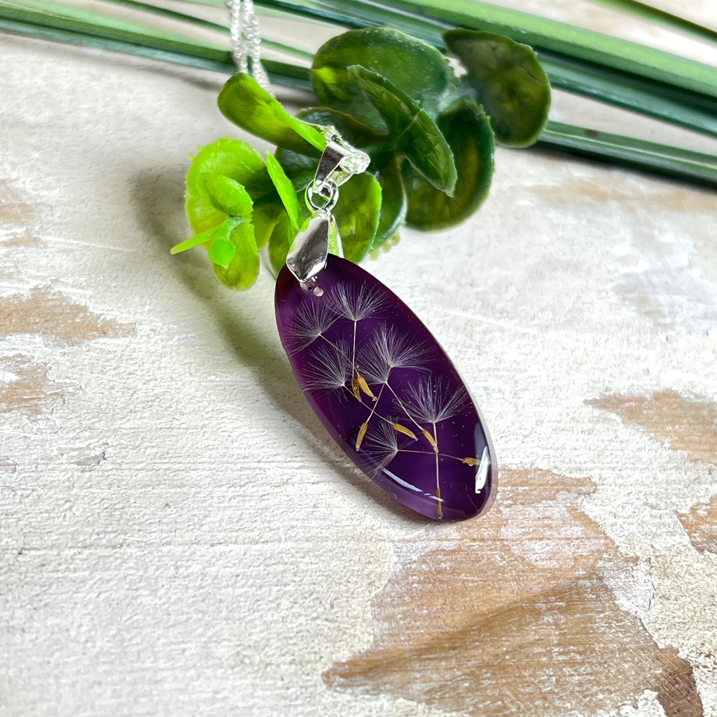 Ovaler Anhänger mit echter Pusteblume in Harz, Hintergrund Brombeerfarben. Größe 4 × 1,8 cm, versilberte Kette 50 cm mit Karabinerverschluss. Handgefertigtes Unikat.