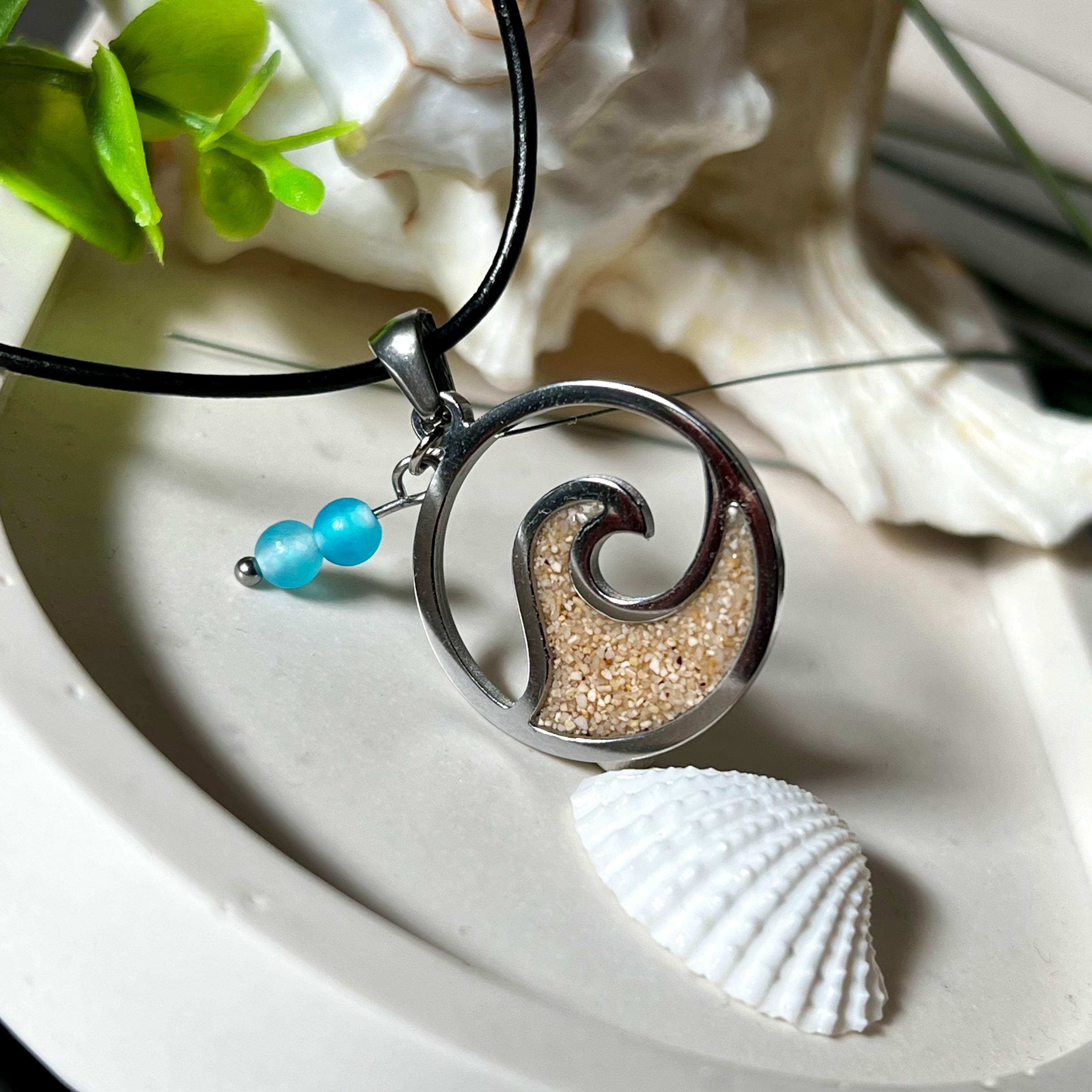 Wave Spirit – runder Edelstahlanhänger mit echter Sandfüllung in Harz eingebettet, inspiriert von einer stilisierten Welle. Verziert mit zwei blauen Achatperlen. Der Anhänger (ca. 2,5 cm Durchmesser) hängt an einer 45 cm langen Lederkette mit Edelstahlverschluss und 5 cm Verlängerungskette. Maritimer Look trifft moderne Eleganz – ein handgefertigtes Schmuckstück für Meerliebhaber