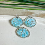 Runder Anhänger „Atlantis Dream“ aus antik-silberfarbener Zinklegierung, gefüllt mit kleinen Larimar-Edelsteinchen und klarem Resin. Die Larimarsteine zeigen sanfte Blau- und Weißtöne, die an das Meer erinnern. Durchmesser ca. 22 mm, handgefertigtes Unikat, fotografiert auf maritimem Untergrund mit kleinen Larimarsteinen und Muscheln zur Größenorientierung.