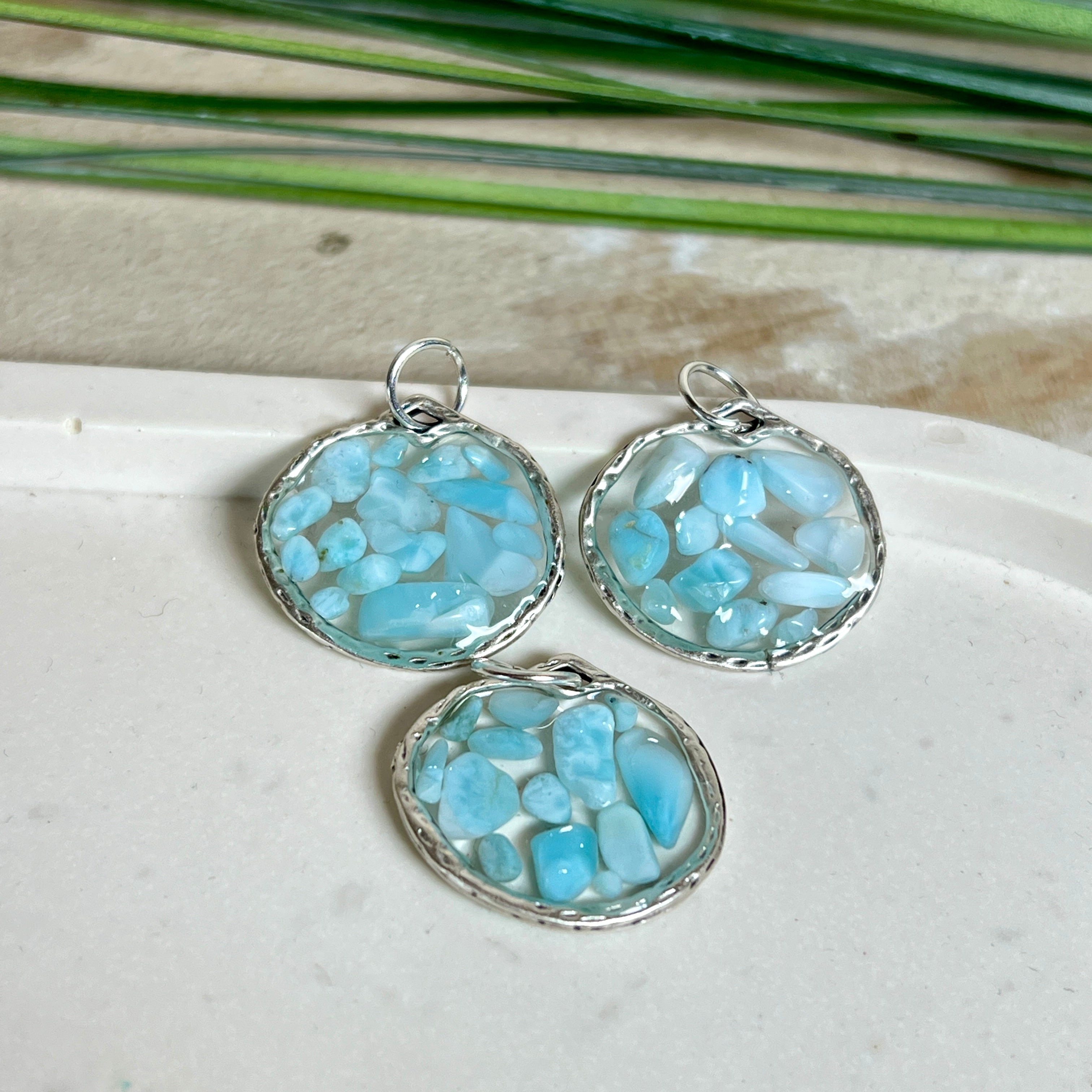 Runder Anhänger „Atlantis Dream“ aus antik-silberfarbener Zinklegierung, gefüllt mit kleinen Larimar-Edelsteinchen und klarem Resin. Die Larimarsteine zeigen sanfte Blau- und Weißtöne, die an das Meer erinnern. Durchmesser ca. 22 mm, handgefertigtes Unikat, fotografiert auf maritimem Untergrund mit kleinen Larimarsteinen und Muscheln zur Größenorientierung.