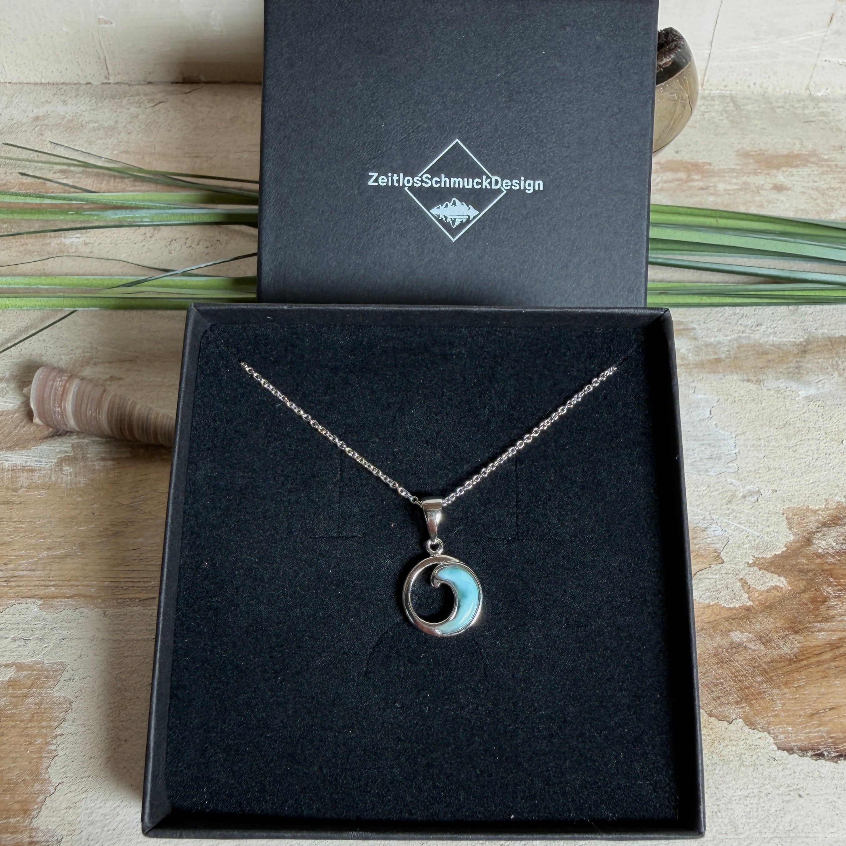 Feine Wellen-Kette aus rhodiniertem 925 Silber. Der runde Anhänger zeigt eine stilisierte Welle mit hellblauem Larimar-Cabochon, eingefasst in poliertem Silber. Größe des Anhängers ca. 23 x 13 mm, an einer 45 cm langen, 1 mm starken, rhodinierten Silberkette. 