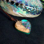 Herzförmiger Anhänger aus Olivenholz mit aquamarinfarbenem Harz und eingelegten Paua-Muschelstücken (Abalone), irisierender Blau-Grün-Schimmer. Der Anhänger hängt an einem braunen, verstellbaren Lederband mit Schiebeknoten; Aufhängung aus Edelstahl. Produktfoto vor maritimer Deko/Abalone-Schale.