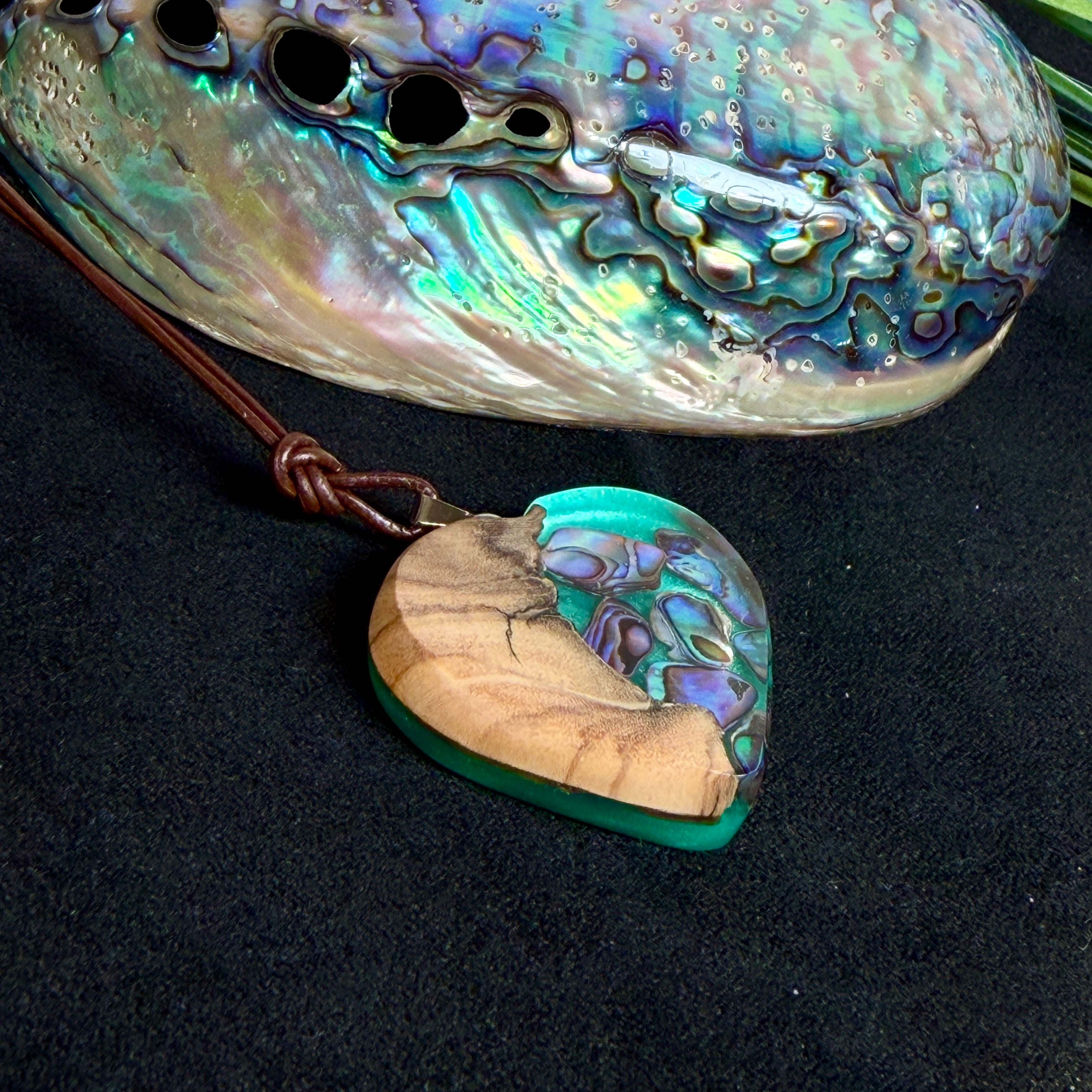 Herzförmiger Anhänger aus Olivenholz mit aquamarinfarbenem Harz und eingelegten Paua-Muschelstücken (Abalone), irisierender Blau-Grün-Schimmer. Der Anhänger hängt an einem braunen, verstellbaren Lederband mit Schiebeknoten; Aufhängung aus Edelstahl. Produktfoto vor maritimer Deko/Abalone-Schale.
