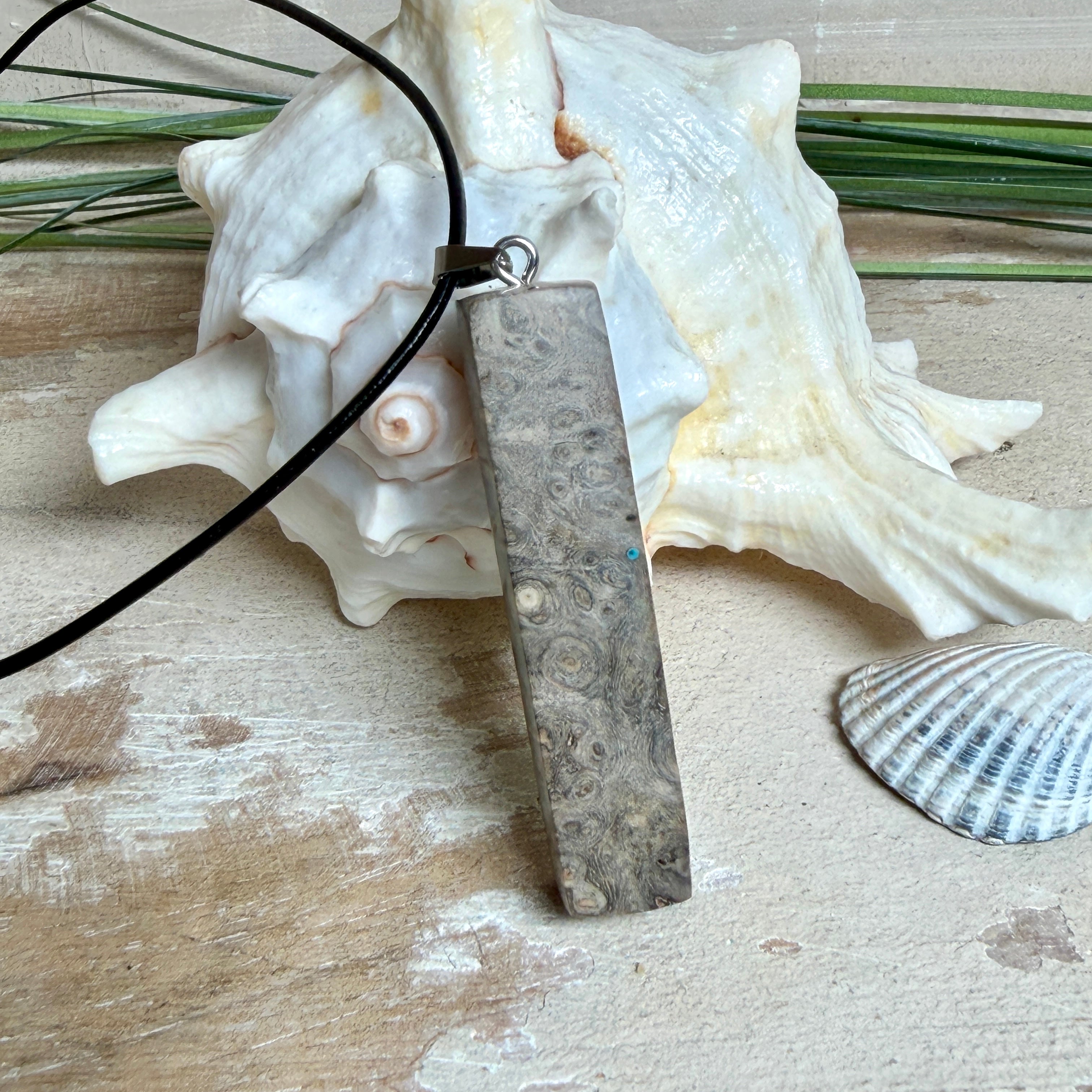 Rechteckiger Stabanhänger aus Maserholz und glasklarem Epoxidharz in tiefen Blau- und Türkistönen mit organischer Lagunen/Riff-Optik. Hochglanzpoliert, ca. 5,3 × 1,2 cm. Der Anhänger hängt an einer schwarzen Lederkette mit Edelstahl-Karabiner und 5-cm-Verlängerung (45 + 5 cm); die Anhängerschlaufe ist ebenfalls aus Edelstahl. Handgefertigtes Unikat, Lieferung in edler Schmuckbox – maritimer Look.