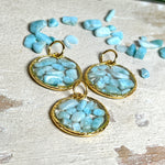 Runder Anhänger „Golden Atlantis Dream“ mit kleinen Larimar-Edelsteinchen in klarem Resin, gefasst in einer 24K vergoldeten Zinklegierung in Shiny Gold. Die Larimarsteine zeigen sanfte Blau- und Weißtöne wie karibische Wellen. Durchmesser ca. 22 mm, handgefertigtes Unikat, fotografiert mit Larimarsteinchen, Muscheln und einer 2-Euro-Münze zur Größenorientierung auf maritimem Untergrund.