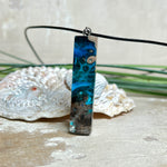 Rechteckiger Stabanhänger aus Maserholz und glasklarem Epoxidharz in tiefen Blau- und Türkistönen mit organischer Lagunen/Riff-Optik. Hochglanzpoliert, ca. 4,6 × 1,2 cm. Der Anhänger hängt an einer schwarzen Lederkette mit Edelstahl-Karabiner und 5-cm-Verlängerung (45 + 5 cm); die Anhängerschlaufe ist ebenfalls aus Edelstahl. Handgefertigtes Unikat, Lieferung in edler Schmuckbox – maritimer Look.