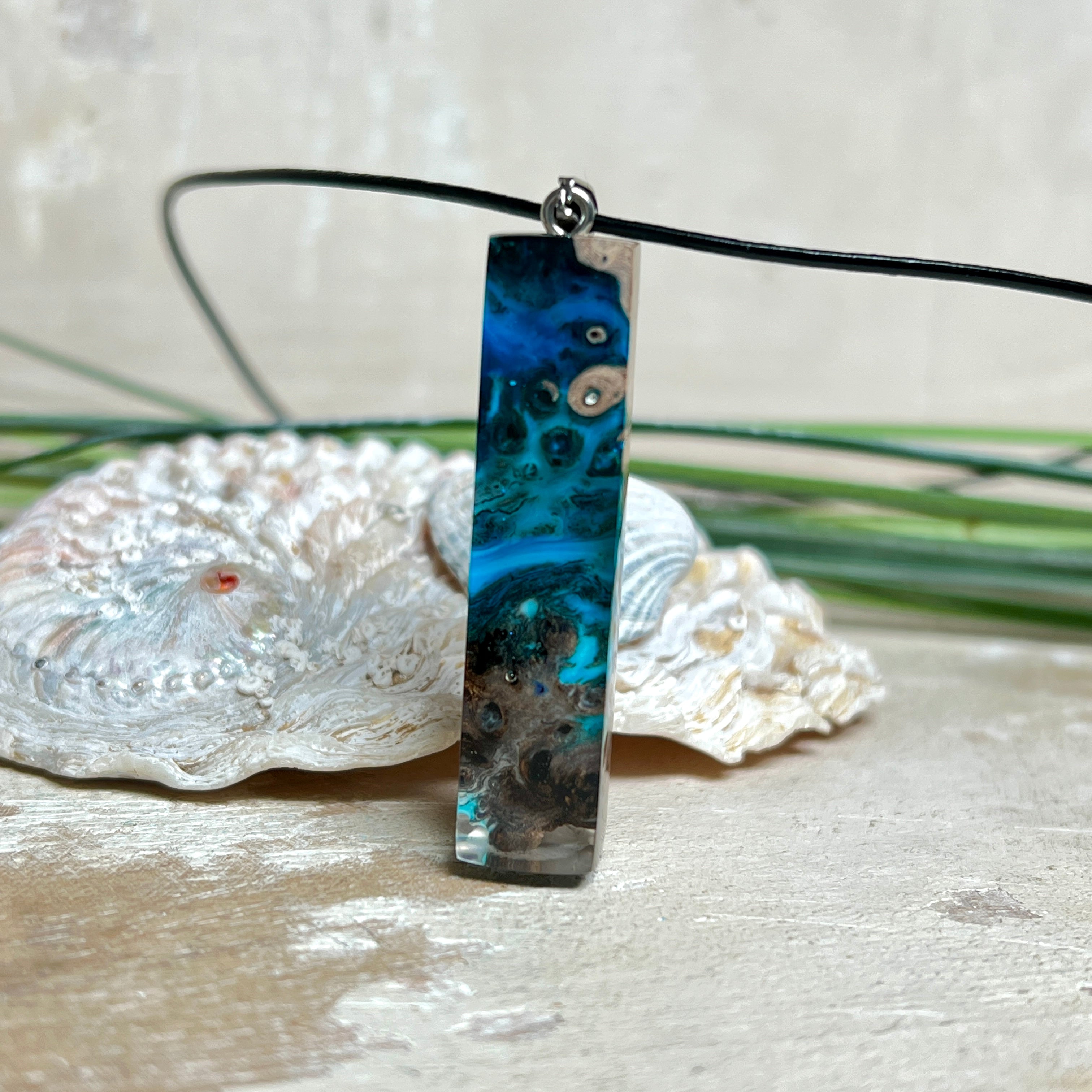 Rechteckiger Stabanhänger aus Maserholz und glasklarem Epoxidharz in tiefen Blau- und Türkistönen mit organischer Lagunen/Riff-Optik. Hochglanzpoliert, ca. 4,6 × 1,2 cm. Der Anhänger hängt an einer schwarzen Lederkette mit Edelstahl-Karabiner und 5-cm-Verlängerung (45 + 5 cm); die Anhängerschlaufe ist ebenfalls aus Edelstahl. Handgefertigtes Unikat, Lieferung in edler Schmuckbox – maritimer Look.