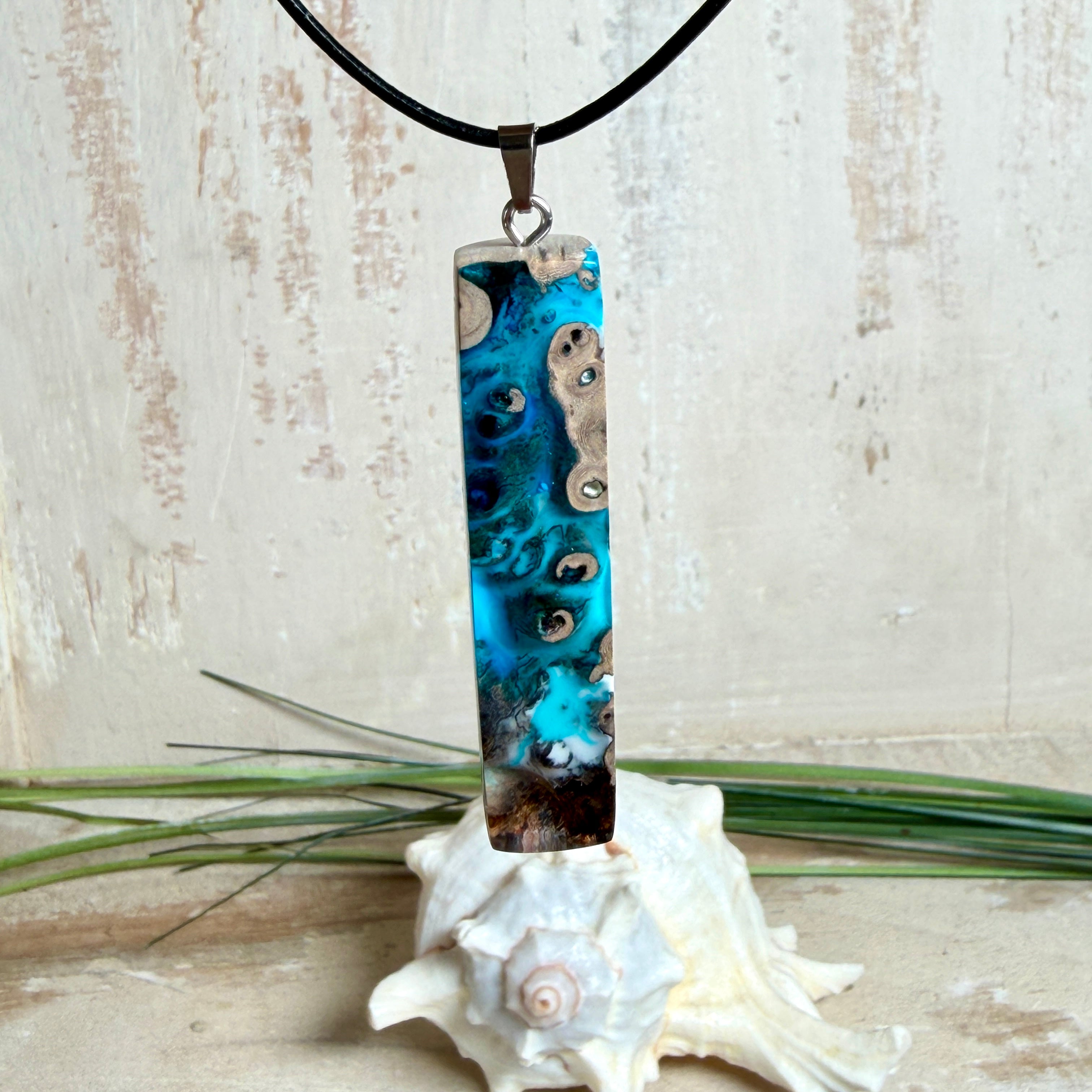 Rechteckiger Stabanhänger aus Maserholz und glasklarem Epoxidharz in tiefen Blau- und Türkistönen mit organischer Lagunen/Riff-Optik. Hochglanzpoliert, ca. 5,3 × 1,2 cm. Der Anhänger hängt an einer schwarzen Lederkette mit Edelstahl-Karabiner und 5-cm-Verlängerung (45 + 5 cm); die Anhängerschlaufe ist ebenfalls aus Edelstahl. Handgefertigtes Unikat, Lieferung in edler Schmuckbox – maritimer Look.