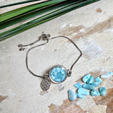 Verstellbares Edelstahlarmband mit Zirkoniasteinchen und Schiebeverschluss und rundem Anhänger (ca. 2,5 × 1,7 cm) mit echten Larimar-Edelstein-Splittern in Harz. Zusätzlich ein Charm „Blume des Lebens“ aus 925 Silber. Handgefertigtes Unikat in Türkis- und Meeresblautönen – Schmuckstück in Nahaufnahme bzw. als Tragebild am Handgelenk.