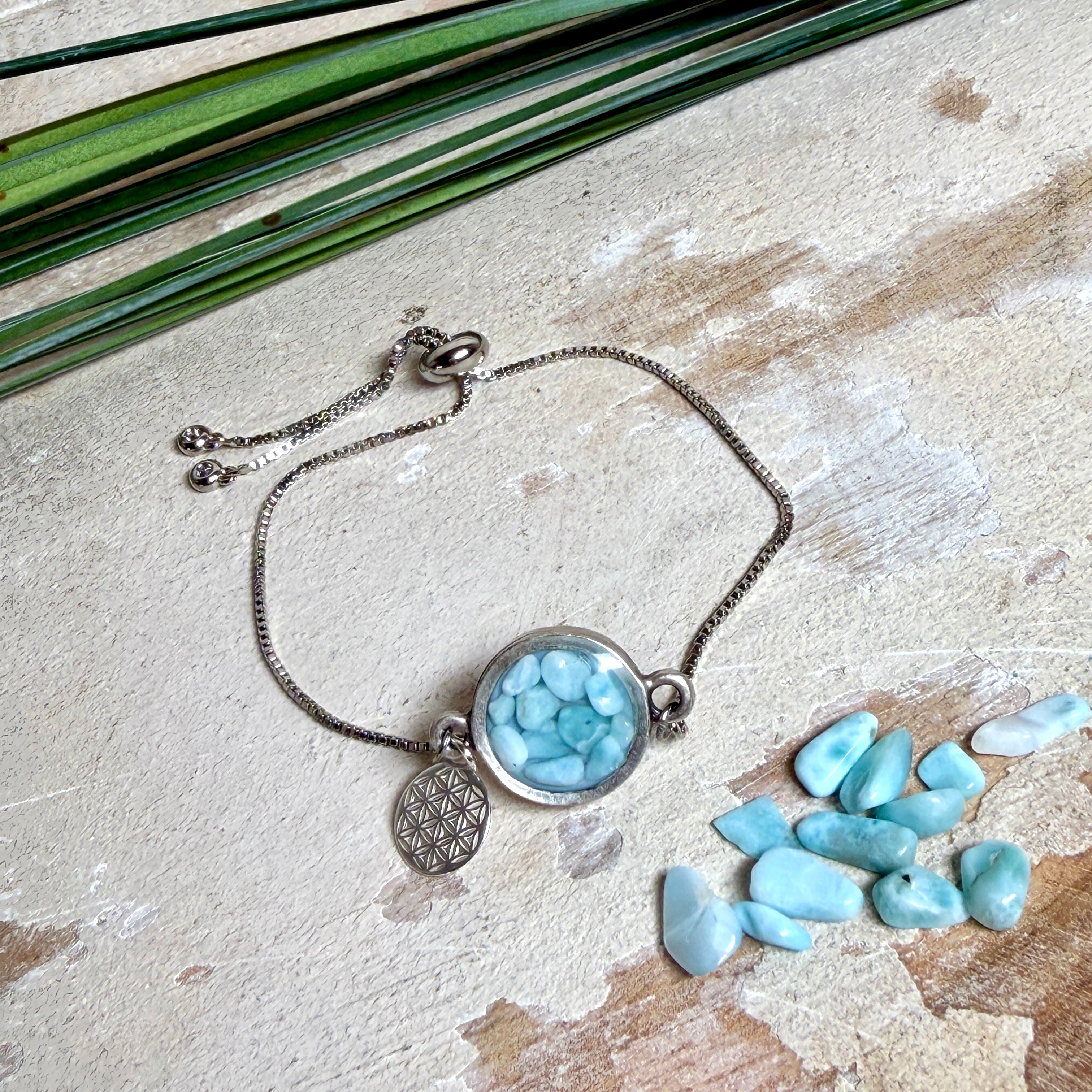Verstellbares Edelstahlarmband mit Zirkoniasteinchen und Schiebeverschluss und rundem Anhänger (ca. 2,5 × 1,7 cm) mit echten Larimar-Edelstein-Splittern in Harz. Zusätzlich ein Charm „Blume des Lebens“ aus 925 Silber. Handgefertigtes Unikat in Türkis- und Meeresblautönen – Schmuckstück in Nahaufnahme bzw. als Tragebild am Handgelenk.