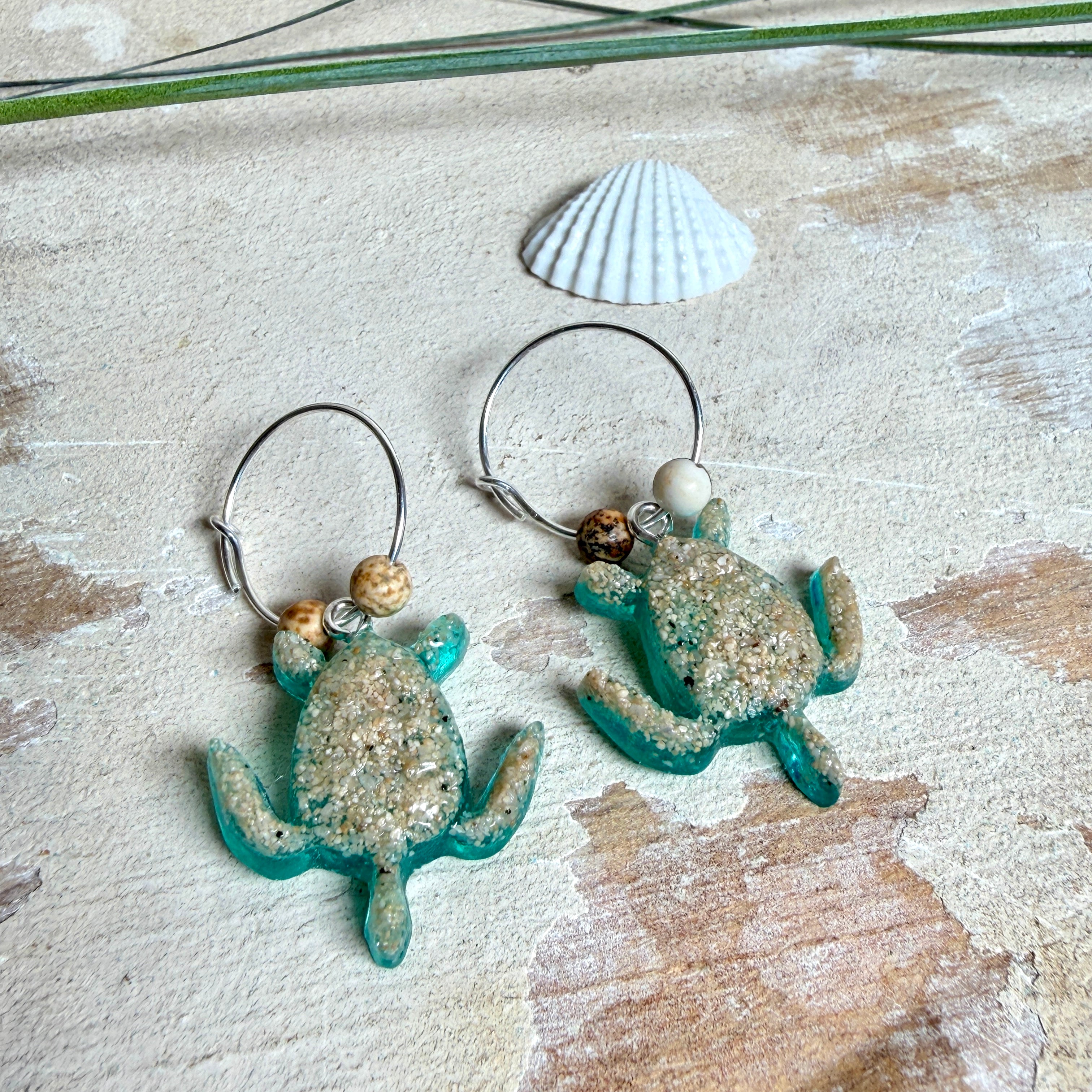 Türkisfarbene Schildkröten-Creolen an feinen 16-mm-Ringen aus 925 Silber, jeweils mit echtem Strandsand im Harz eingebettet und mit kleinen Jaspis-Edelsteinperlen verziert; handgefertigtes Unikat mit sommerlich-maritimem Look, fotografiert als Freisteller, Nahaufnahme oder auf dekorativem Untergrund.