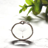 „Handgefertigter Pusteblumen Anhänger mit echter Löwenzahn Pusteblume in klarem Resin, runder silberfarbener Rahmen, filigraner Natur Schmuck auf hellem Marmor Hintergrund.“